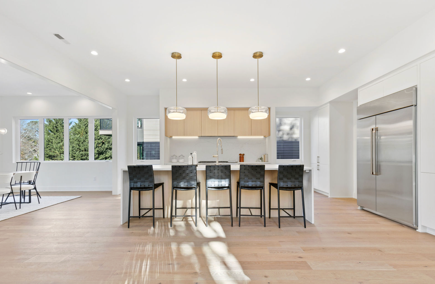 Modern open-concept kitchen with island, bar stools, pendant lights, and stainless steel fridge on light hardwood floors.