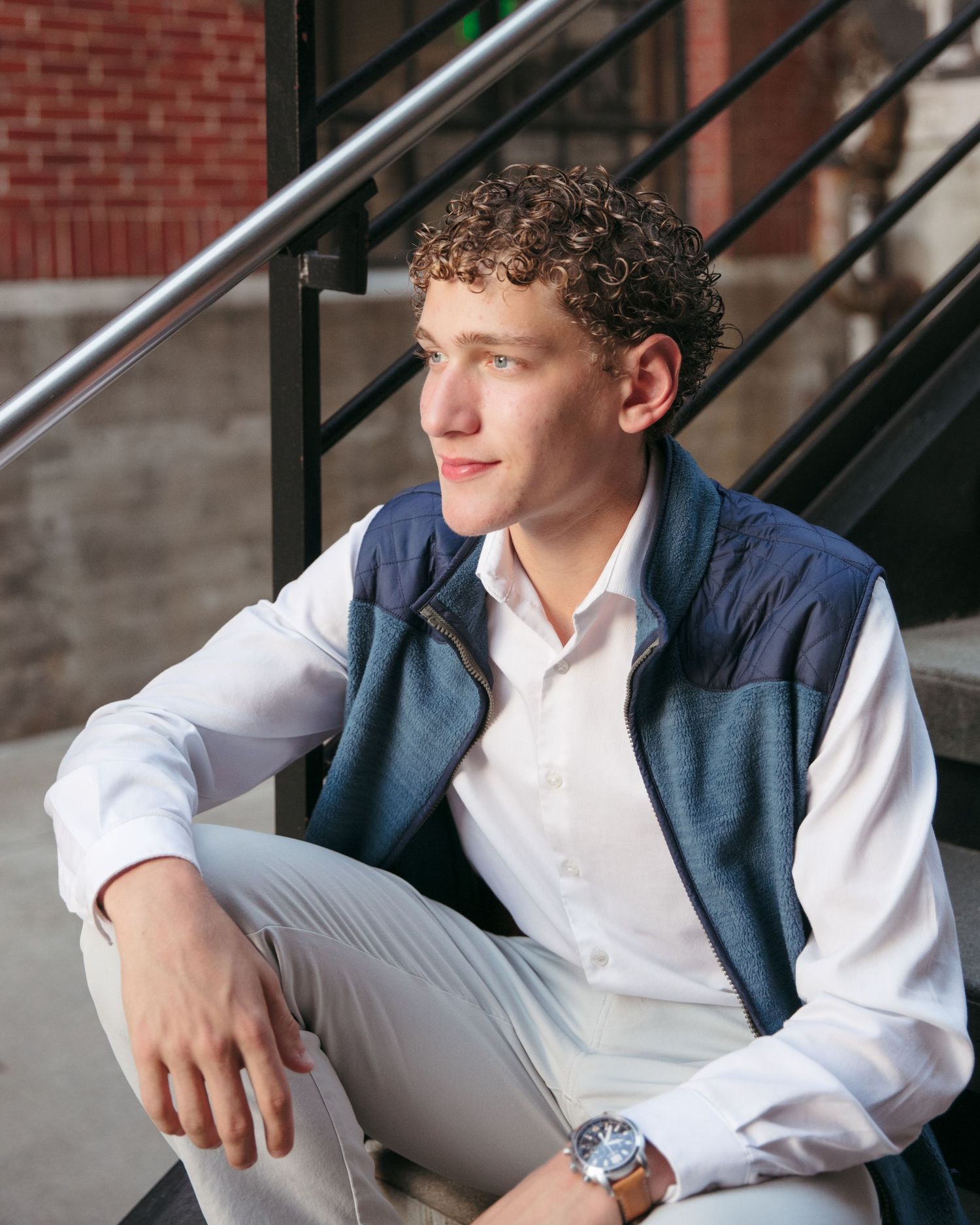 Person sitting on stairs wearing a navy vest and white shirt in an urban setting.