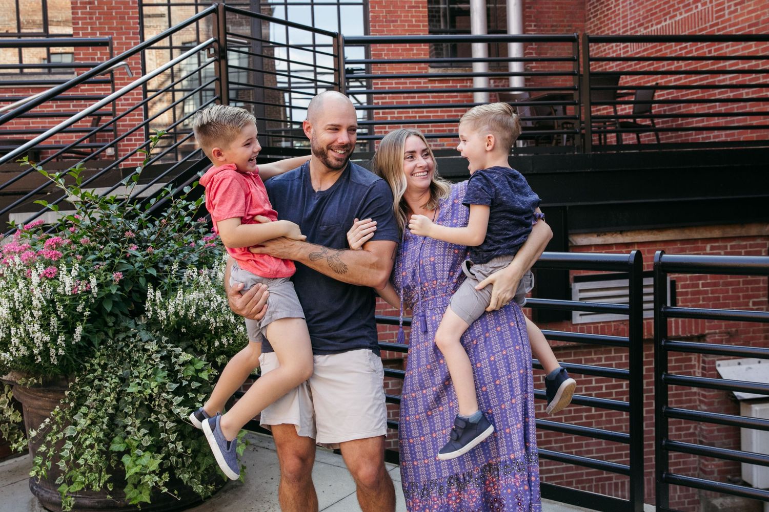 A playful family moment outside a brick building with greenery and metal railings.