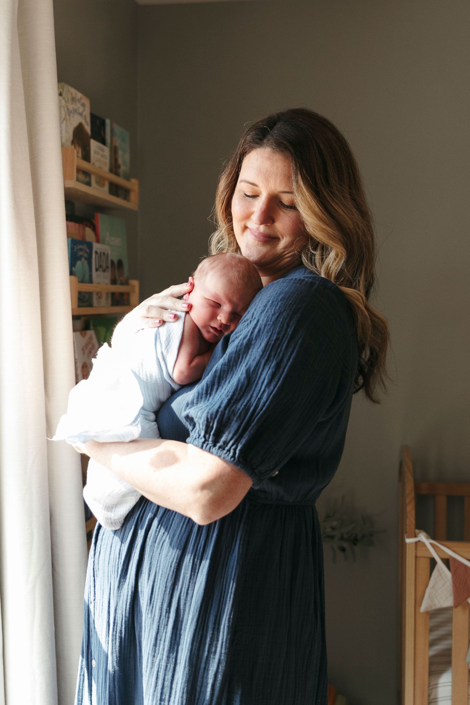 Person in blue dress holding sleeping newborn by sunny window.