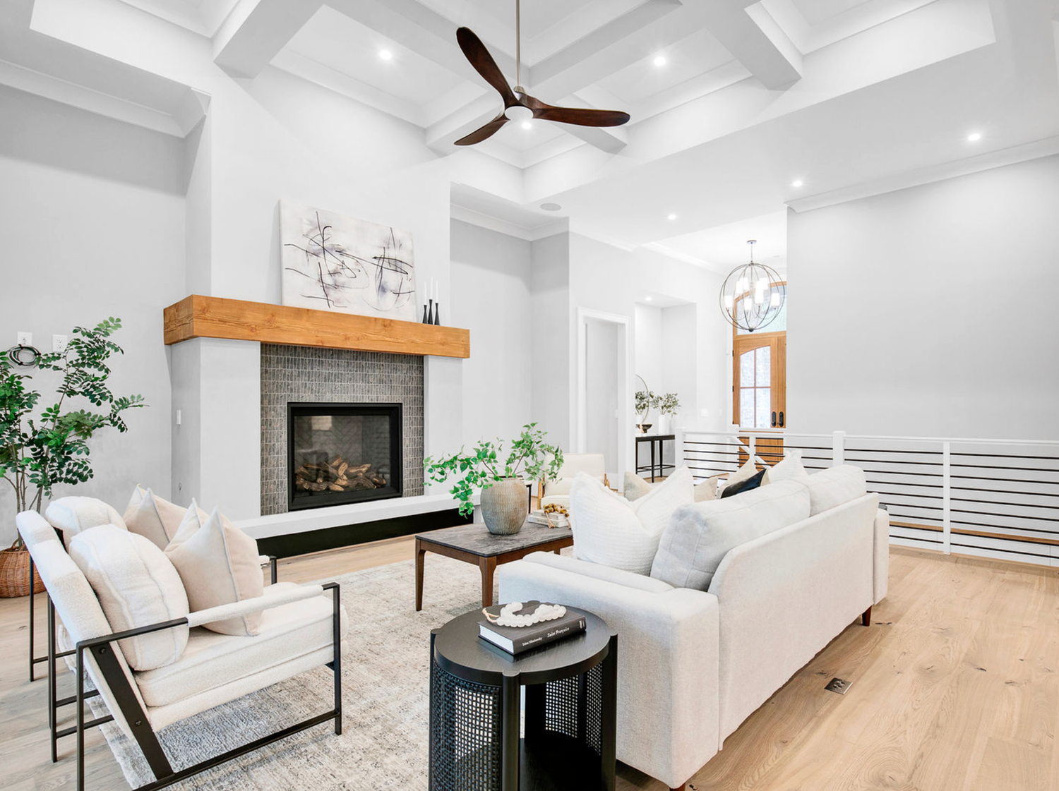 Bright living room with coffered ceiling, wood-beam fireplace, white sofas, ceiling fan, and hardwood floors.