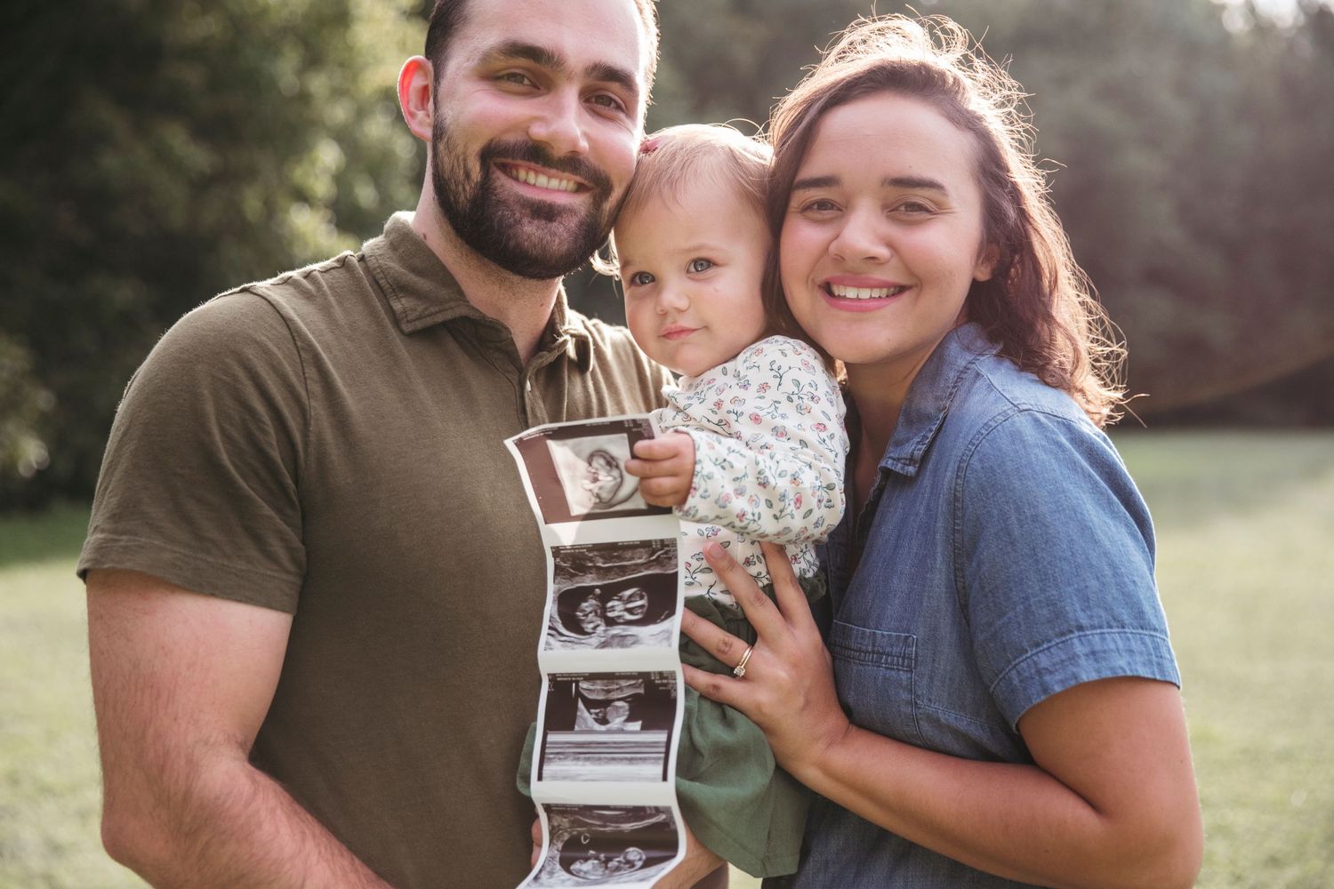 Family holding ultrasound photos while embracing outdoors in a sunny field.
