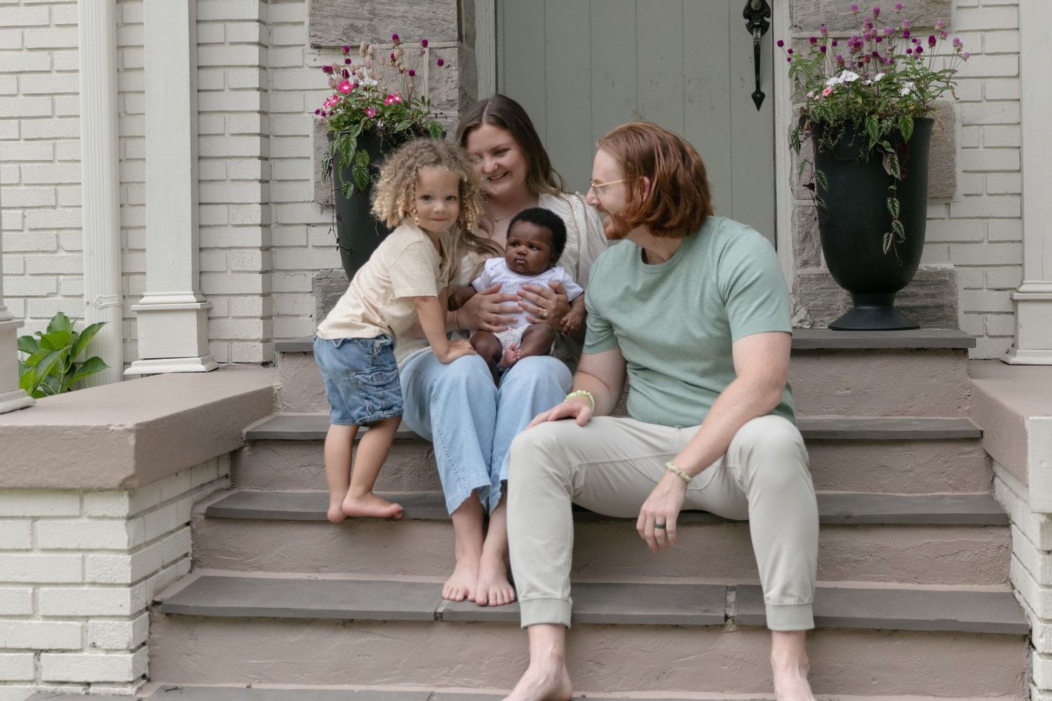 A happy family sits together on white brick porch steps with potted flowers.