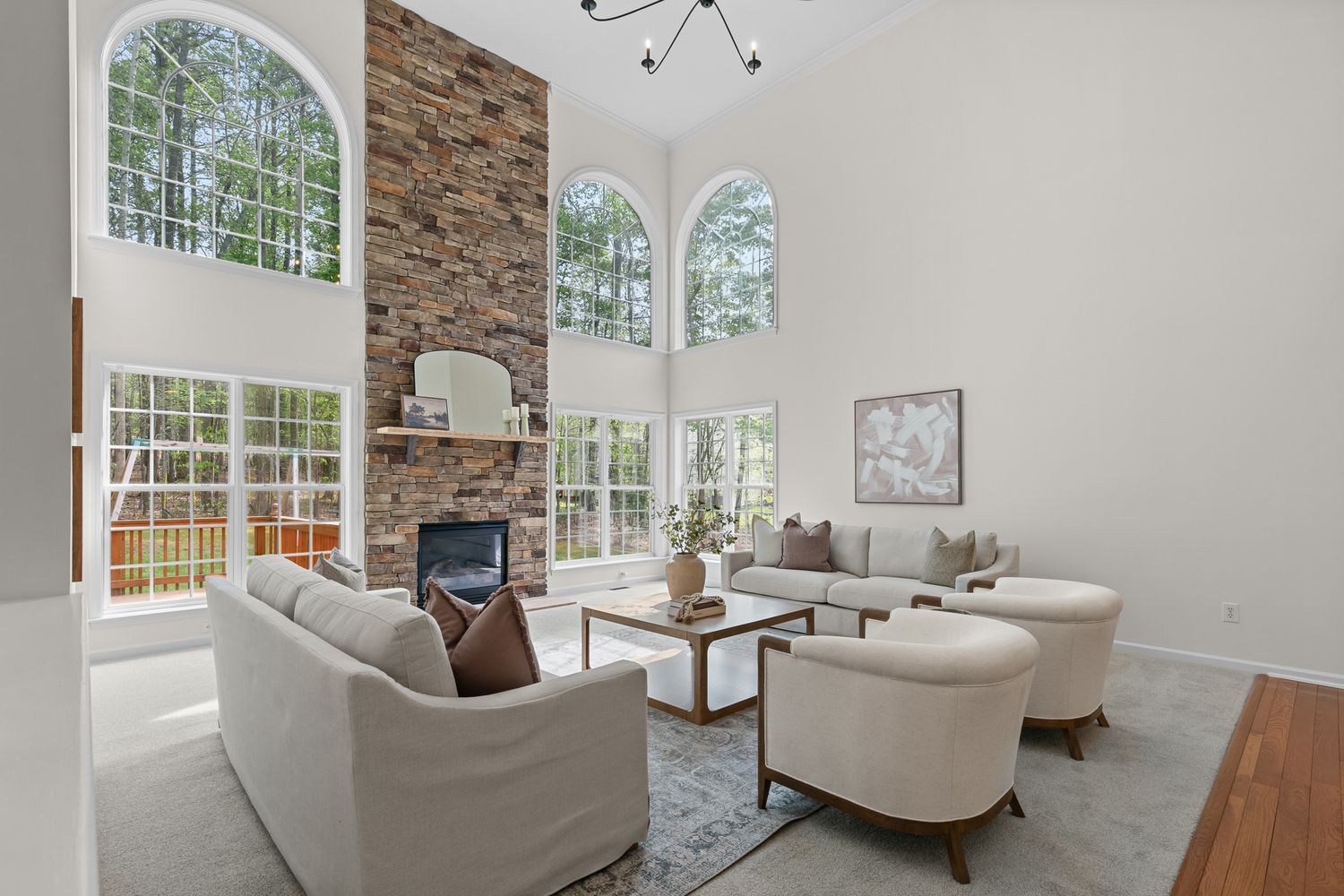 Bright living room with vaulted ceilings, stone fireplace, white sofas, and arched windows overlooking lush greenery.