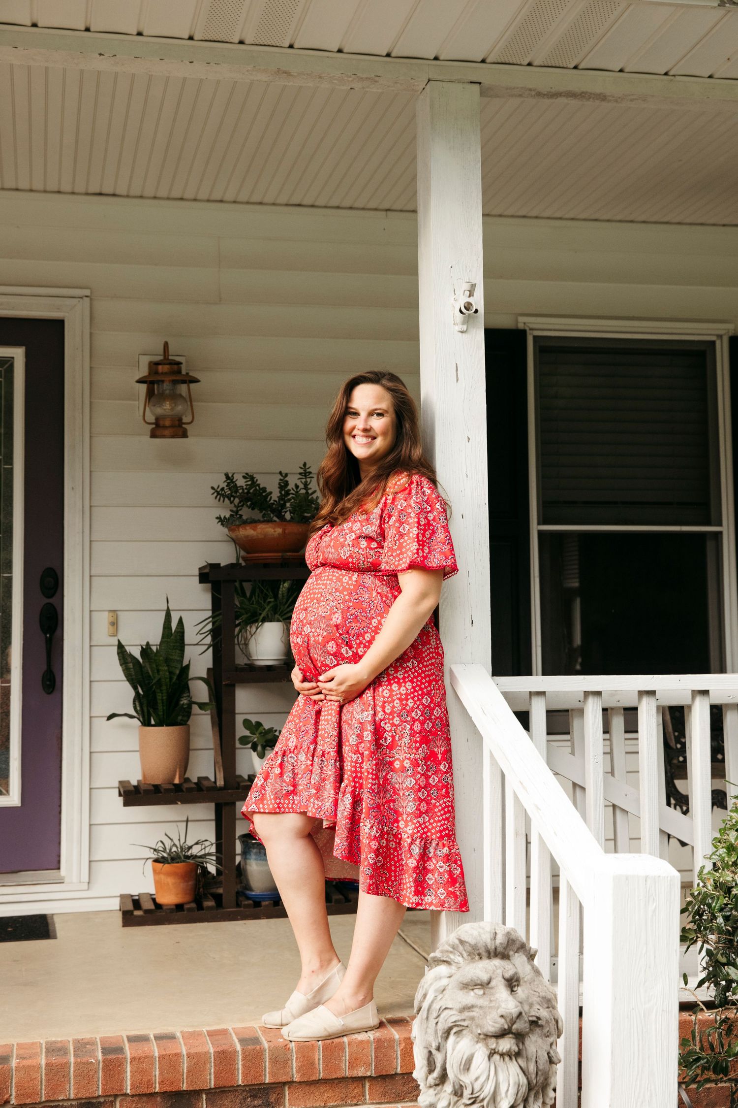Person in red floral dress sitting on white porch railing of traditional farmhouse.