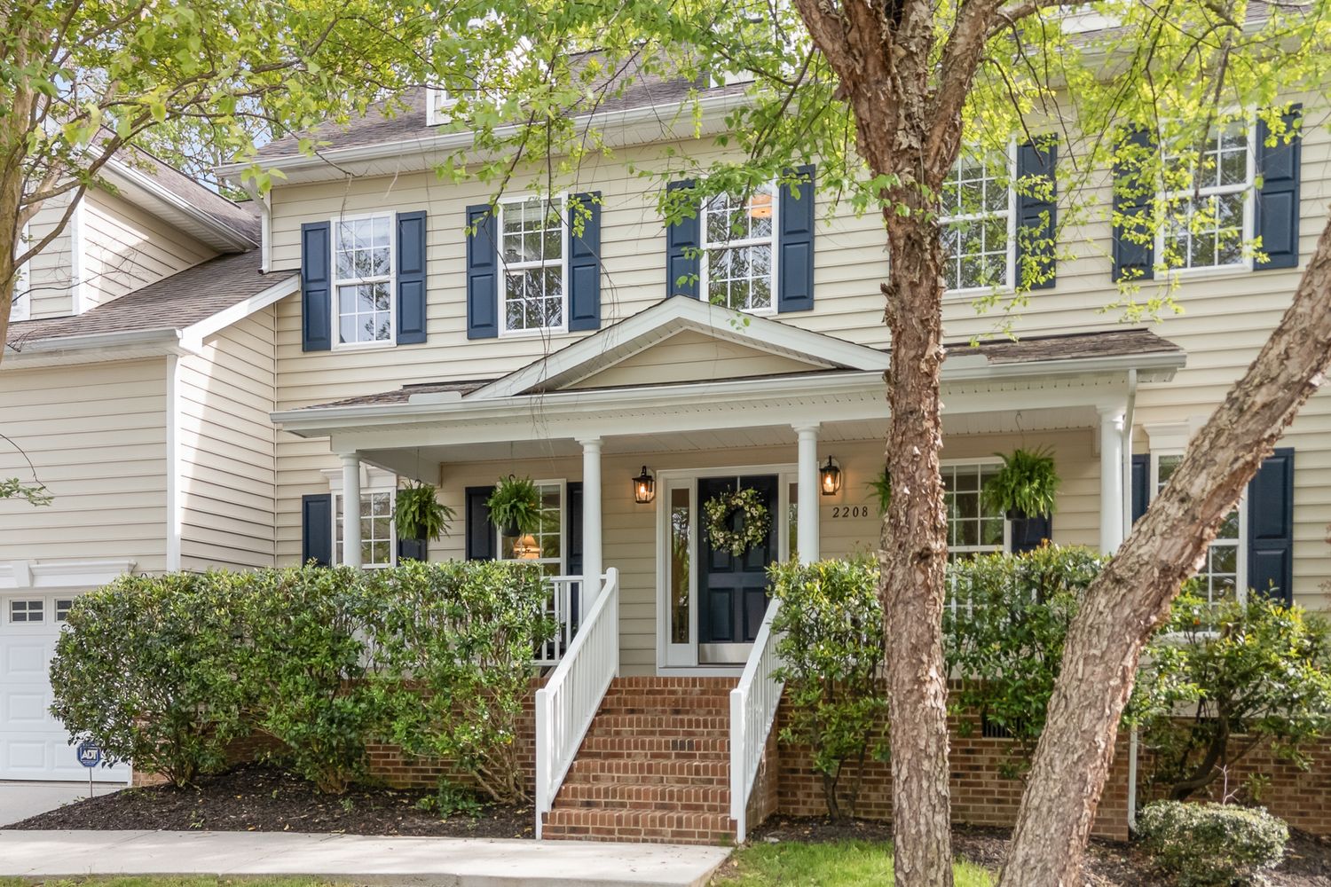 Charming two-story beige home with black shutters, covered front porch, brick steps, and mature trees in a suburban neighborhood.