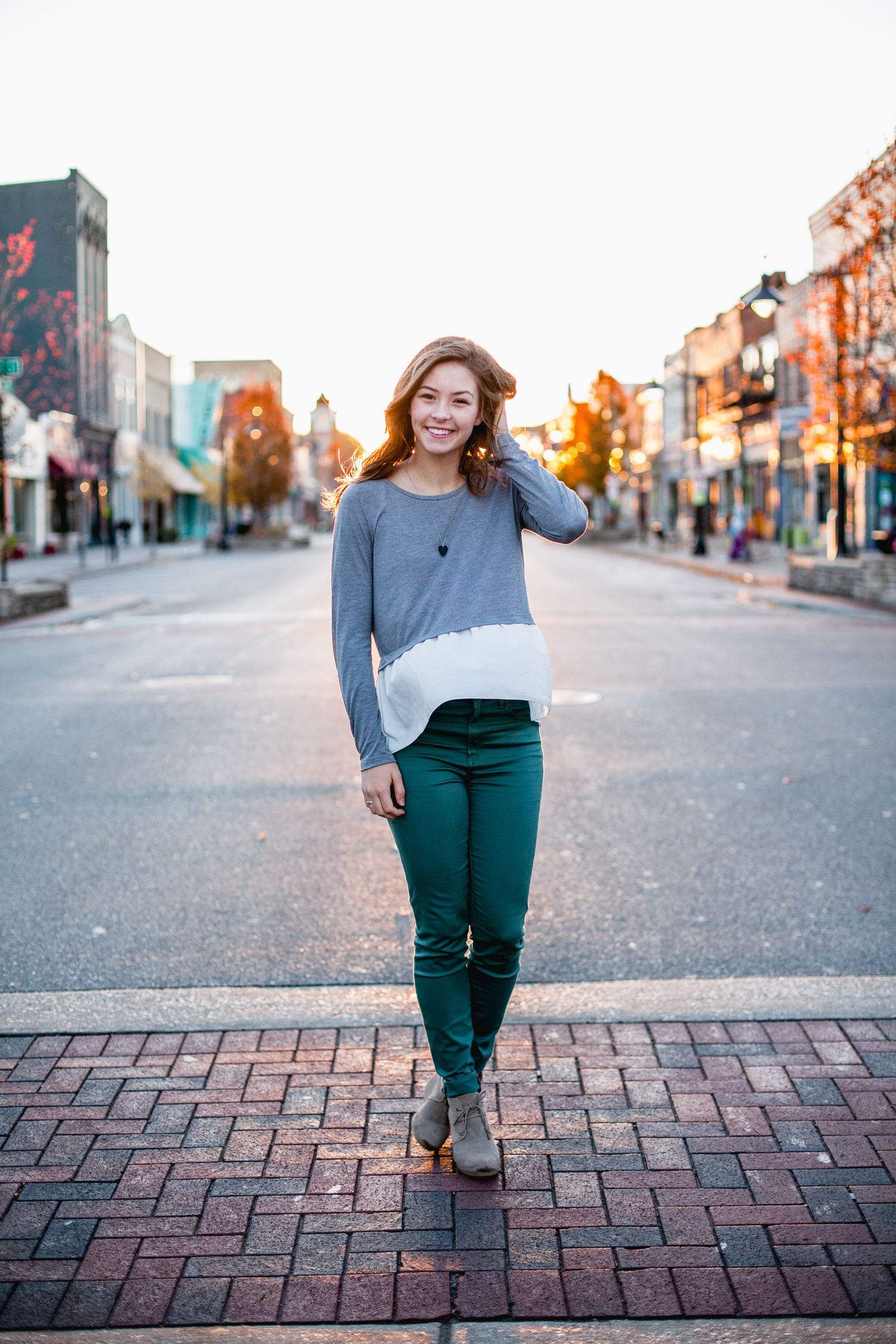 Woman in gray sweater and green pants poses casually on brick sidewalk in autumn urban setting.