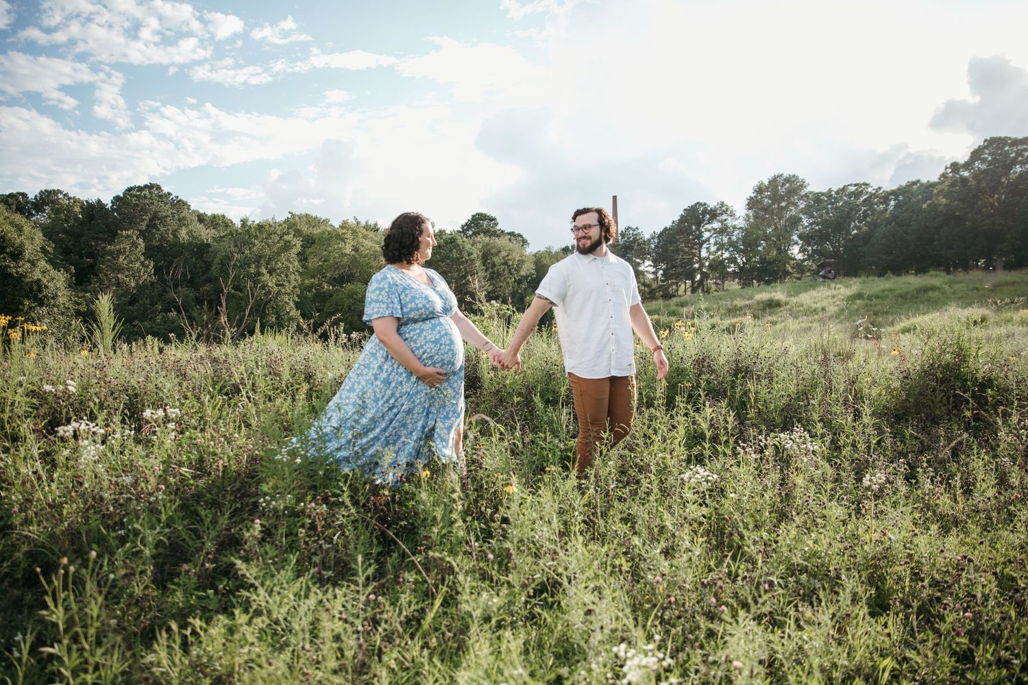 Couple stands among wildflowers in sunny meadow during maternity photoshoot.