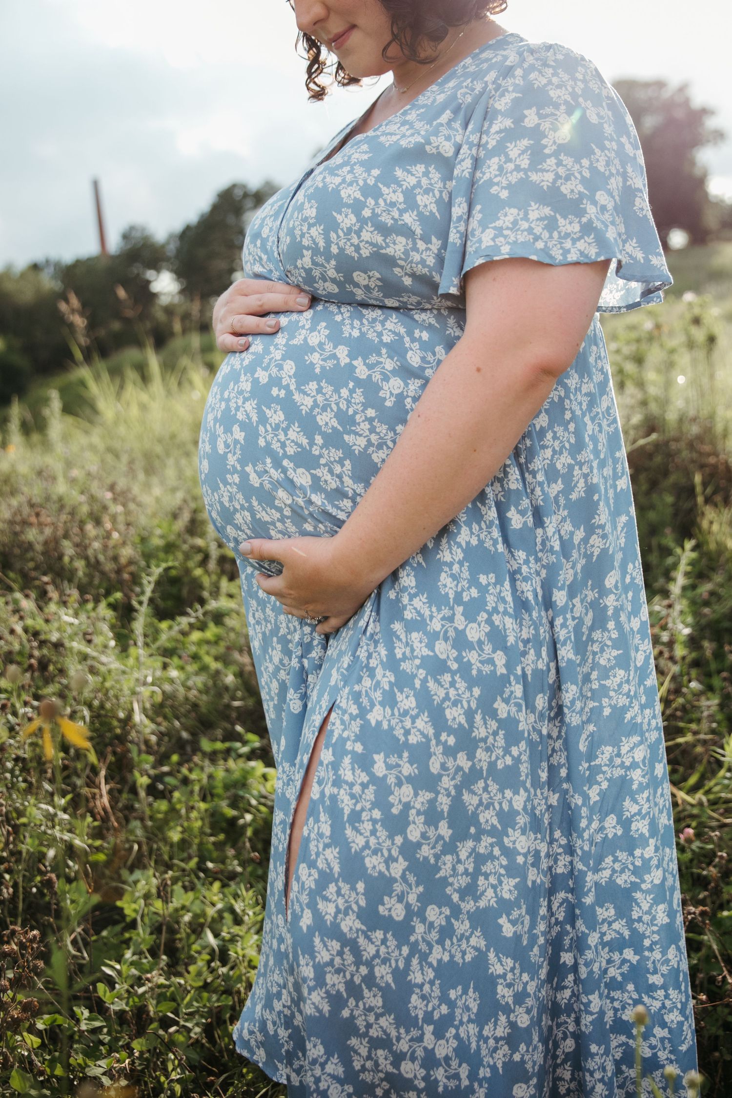 A pregnant person wearing a blue floral dress stands in a sunlit meadow during a maternity photoshoot.