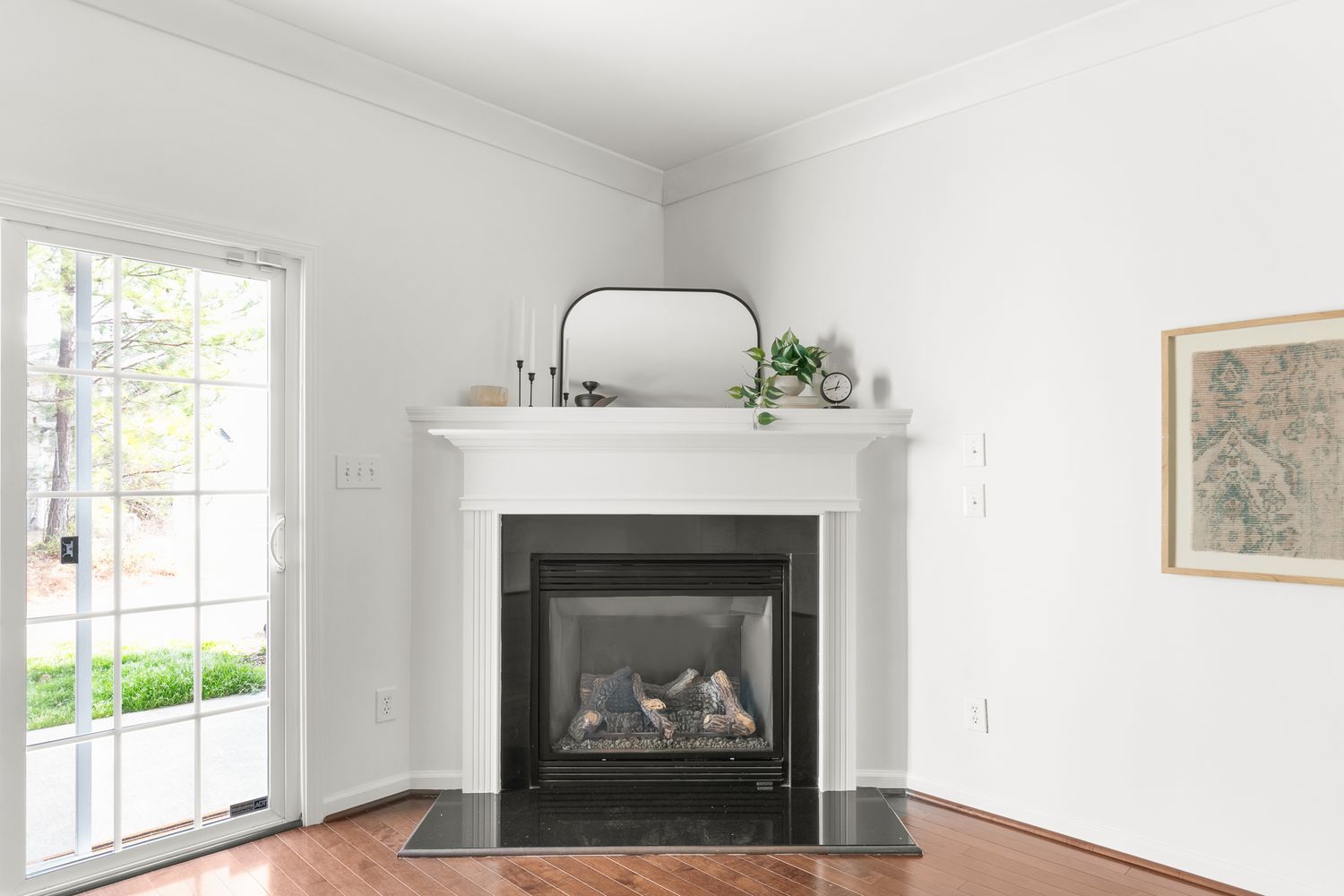 White corner fireplace with black insert, mirror above mantel, hardwood floors, and natural light from adjacent glass door.