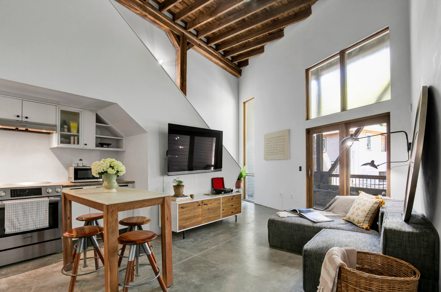 Modern loft living room with exposed wood beam ceiling, concrete floors, open kitchen, and large windows flooding natural light.