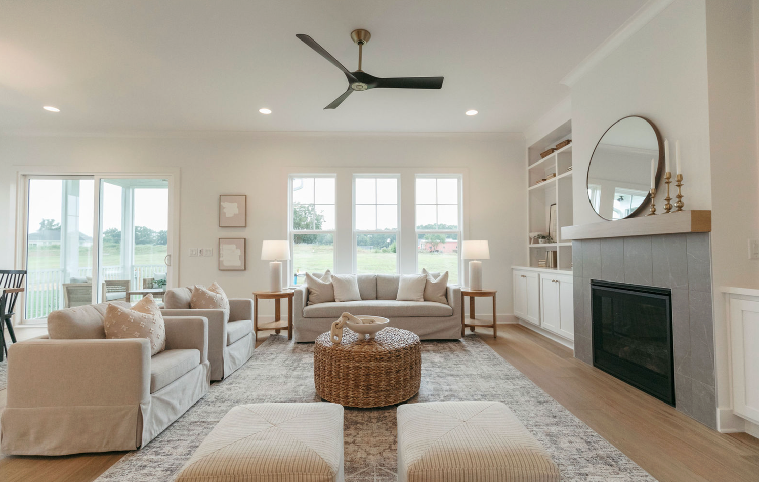 Bright farmhouse living room with beige sofas, wicker ottoman, ceiling fan, built-in shelves, and gray fireplace.