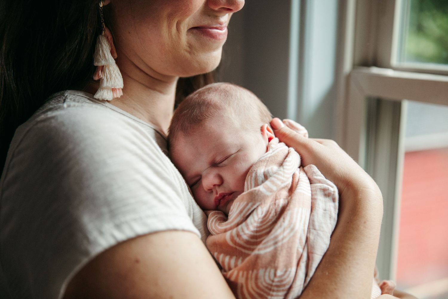Sleeping newborn in pink swaddle rests peacefully against parent by window.
