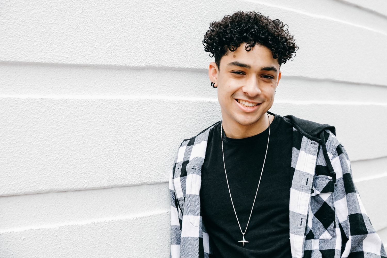 Person wearing plaid shirt and necklace smiles against white wall.