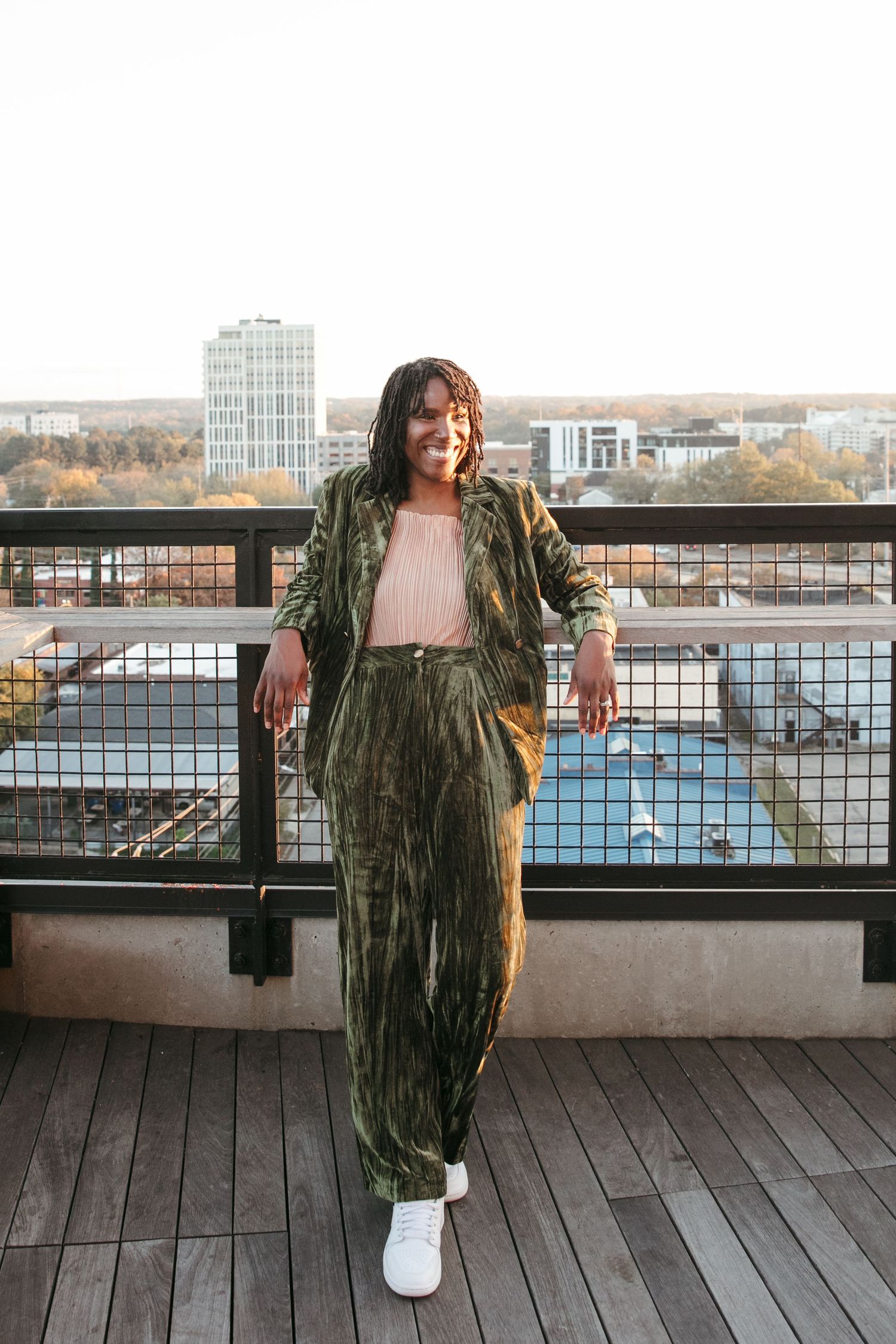 Person in green velvet jacket poses on wooden deck with cityscape and water view in background.