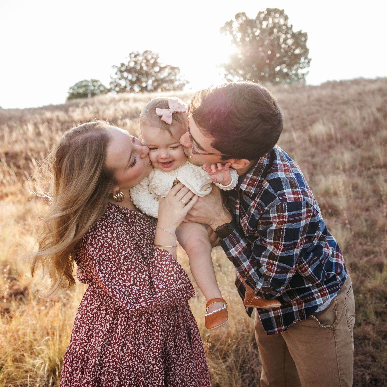 Parents kiss their laughing baby while standing in a sun-lit meadow.