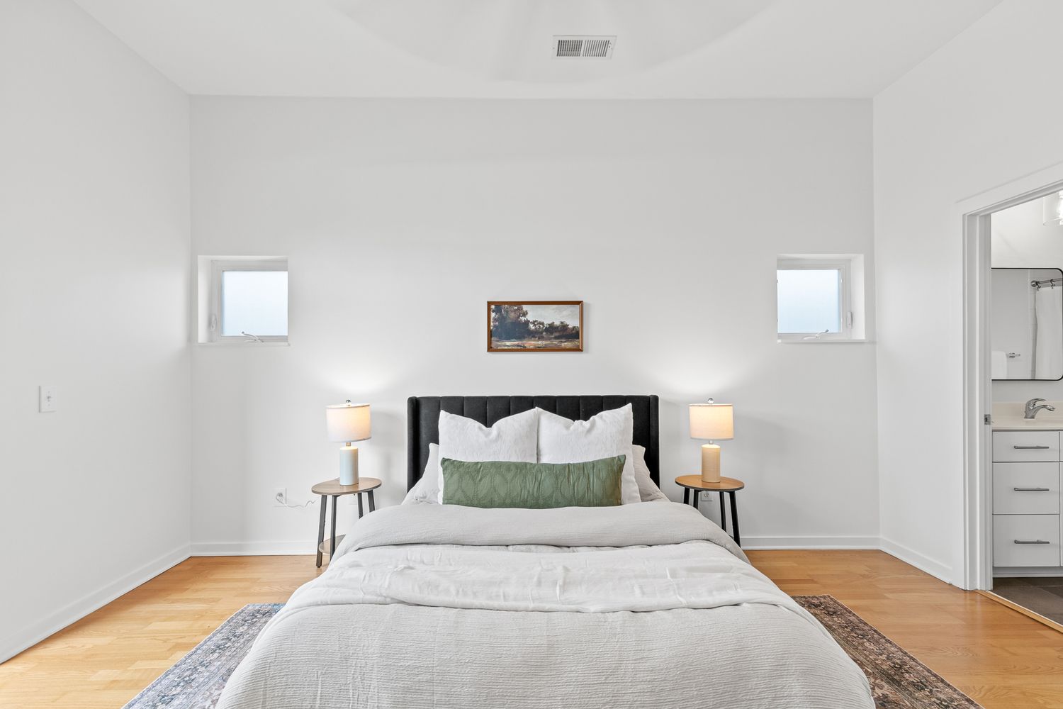 Bright minimalist bedroom with white walls, hardwood floors, queen bed with green pillows, and twin windows above.