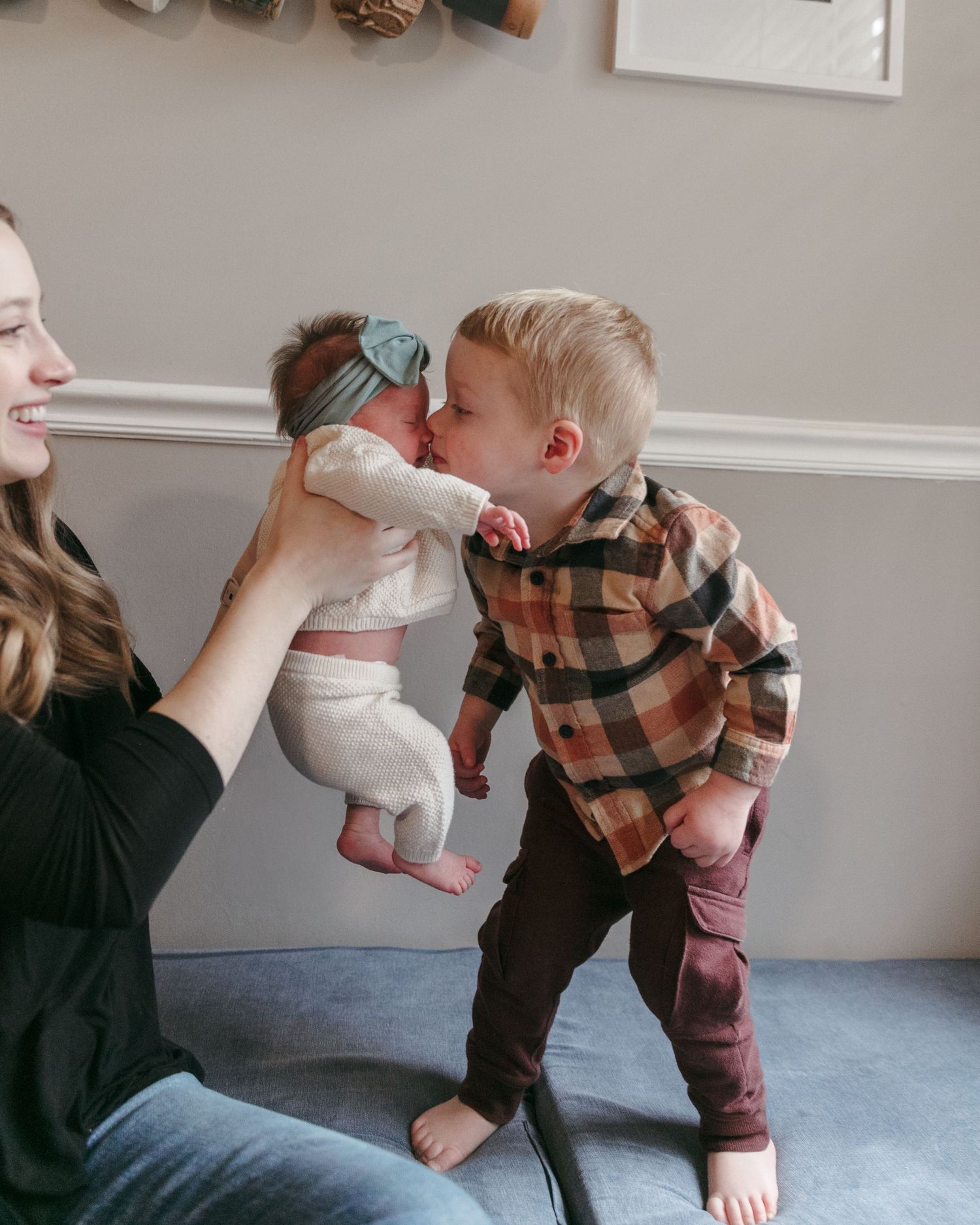 A heartwarming family scene with a baby in white being kissed by a sibling in a plaid shirt.