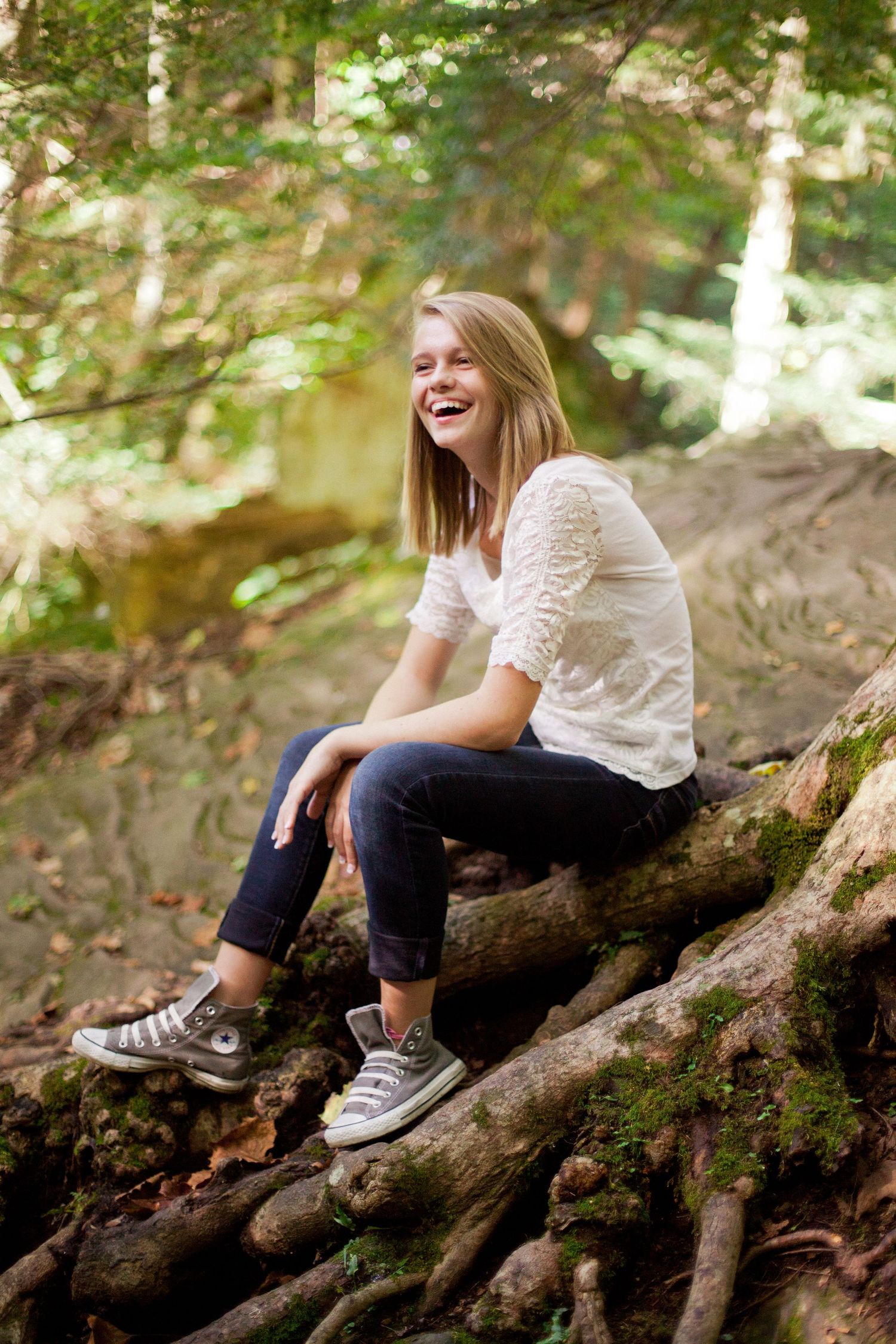 Person in white top and jeans sits laughing on tree roots in forest setting.