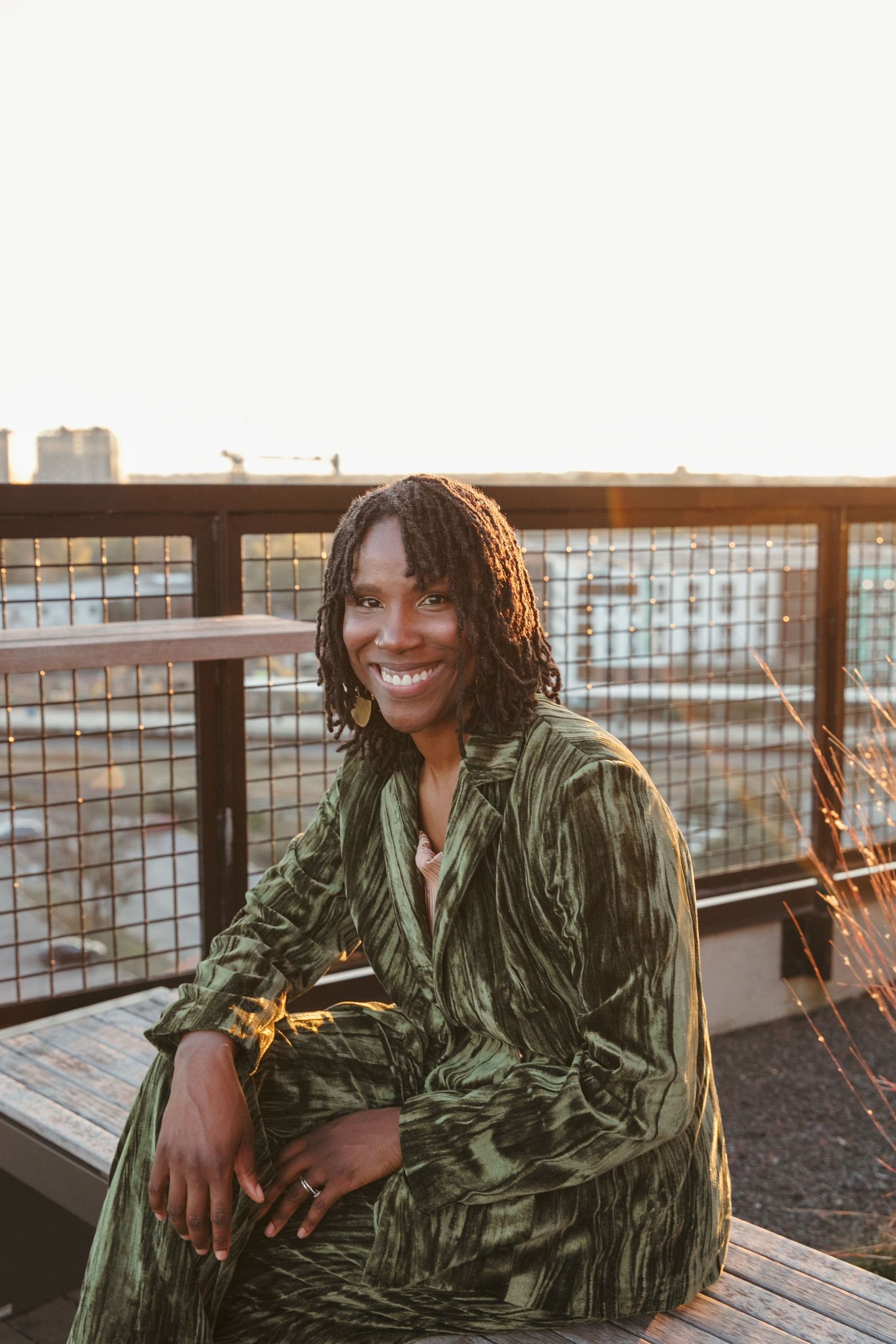 Person in emerald velvet attire poses casually on wooden deck with urban background.