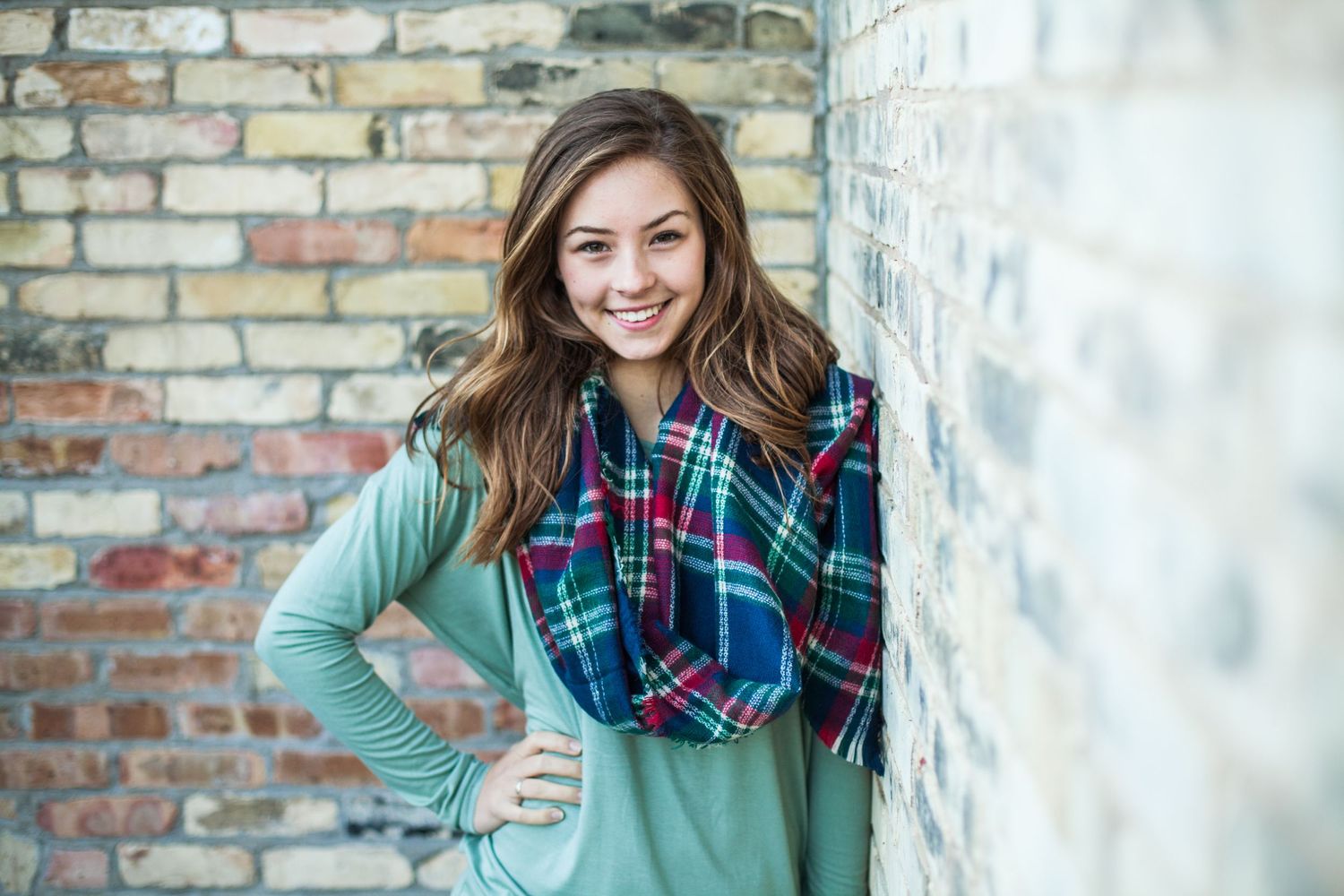 A person in a mint sweater and plaid scarf leans against a brick wall, smiling brightly.