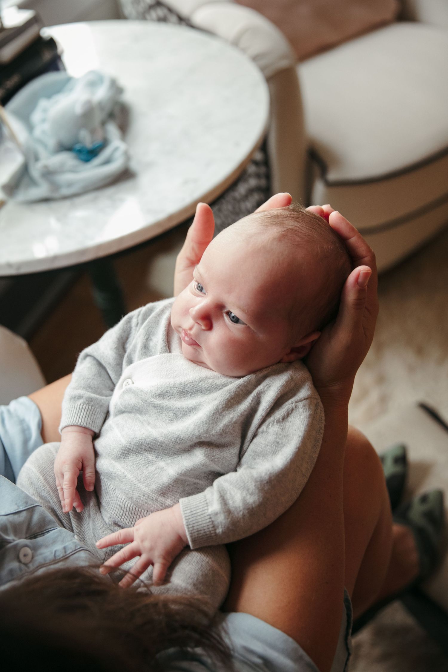 Baby in gray onesie rests in parent's hands on living room couch.