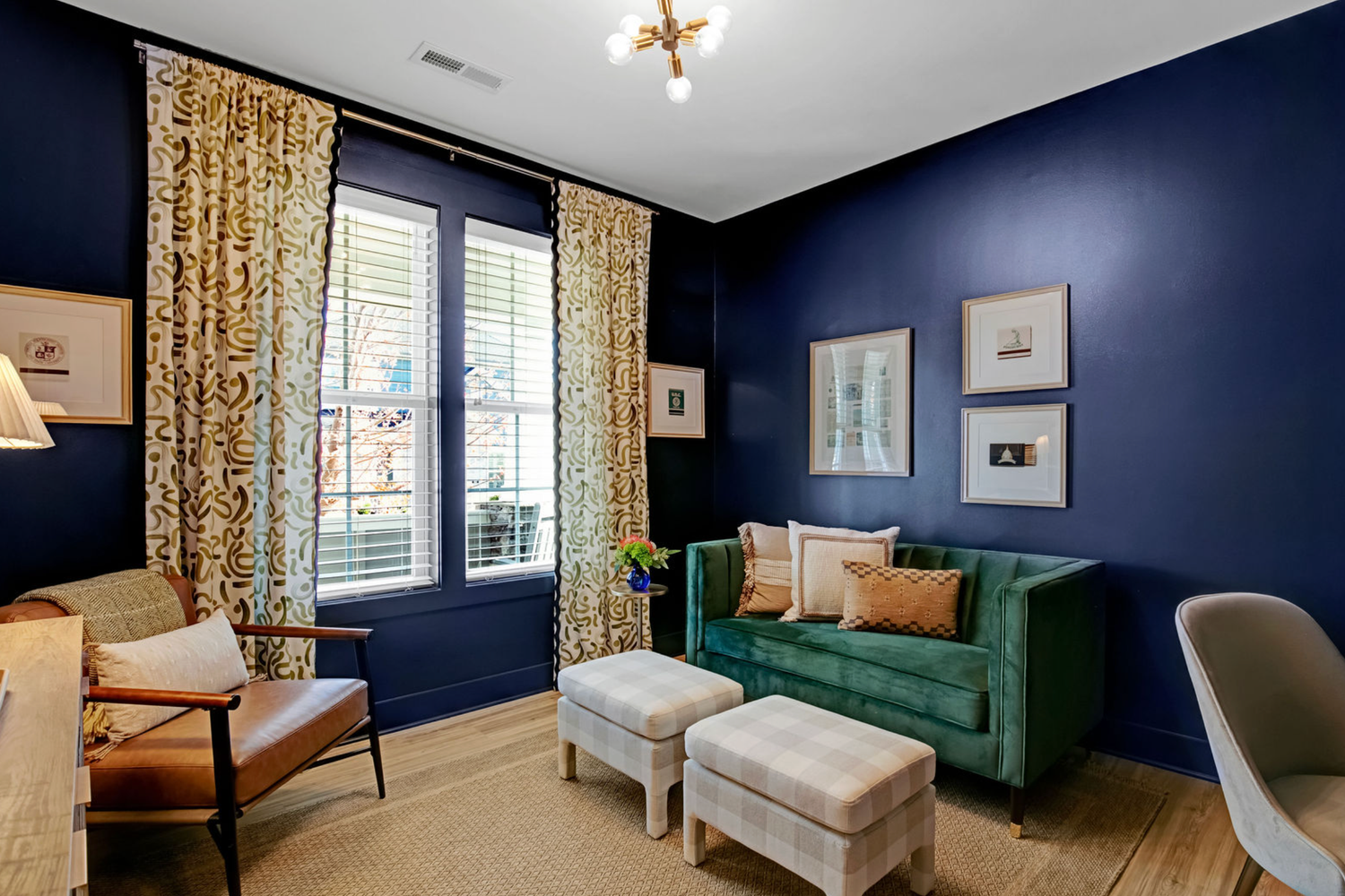 Elegant sitting room with navy blue walls, green velvet sofa, patterned curtains, and neutral ottomans in a modern home.