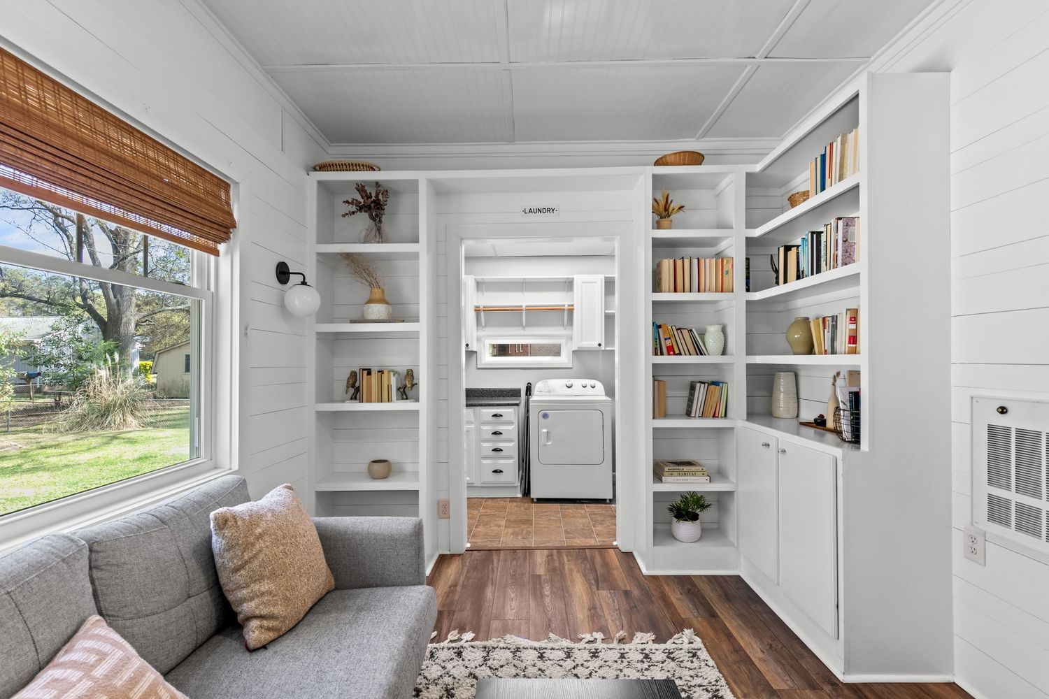Bright tiny home interior with white built-in shelves, gray sofa, hardwood floors, washer, and large window with natural light.