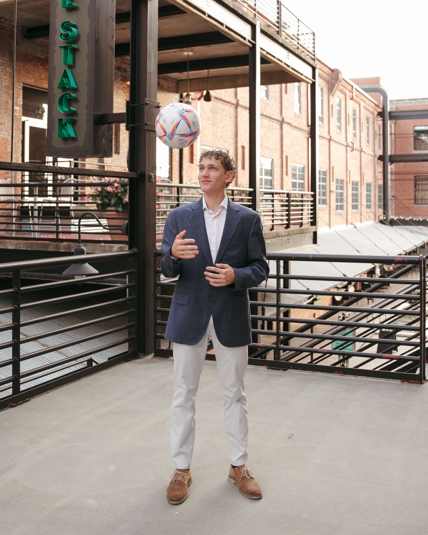 A full-length portrait of someone in business casual attire against an urban brick building setting.