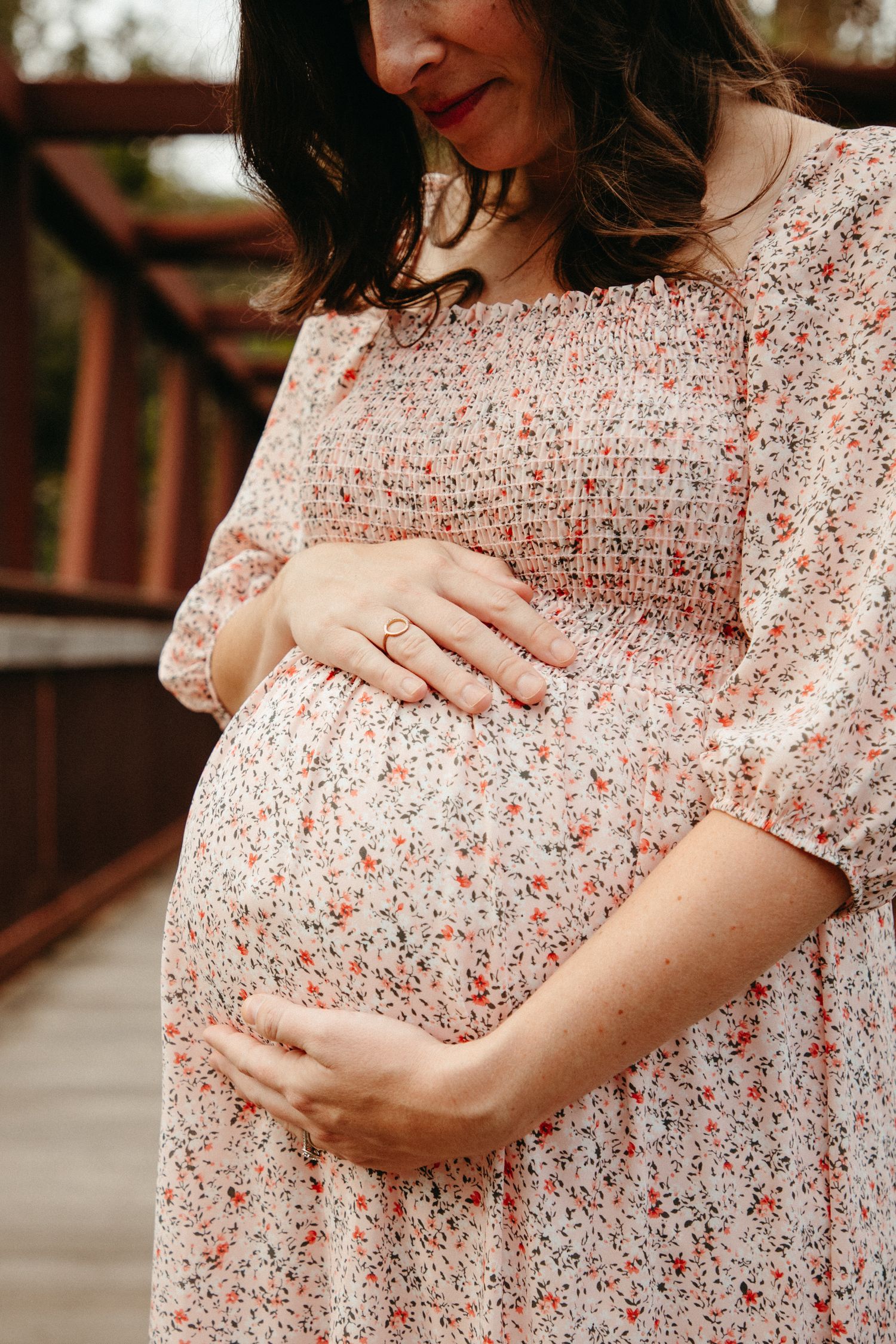 Close-up of a pregnant belly being lovingly held in a floral maternity dress on a rustic bridge.