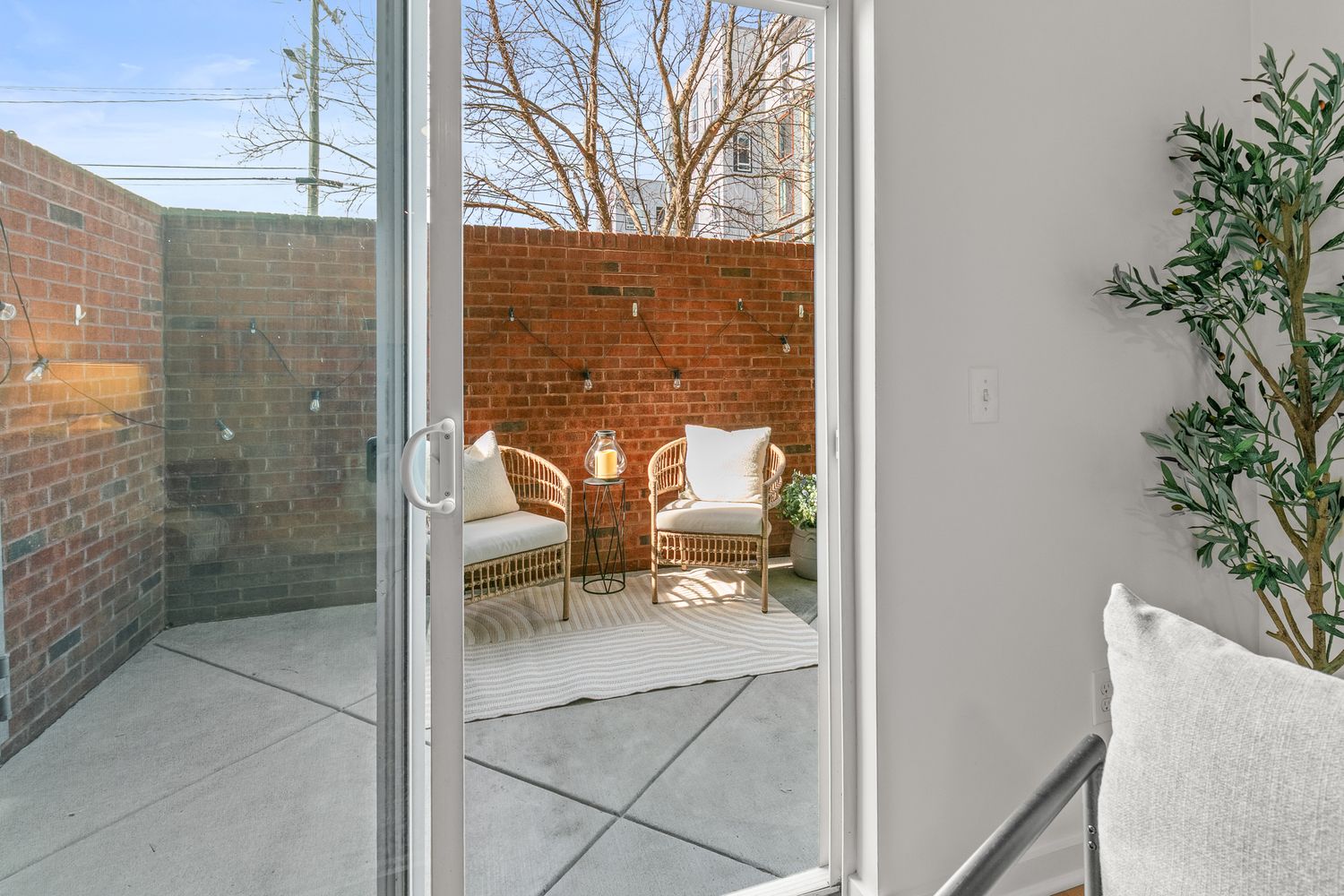 Sliding glass door opening to a cozy brick-walled patio with wicker chairs, cushions, and a small side table.