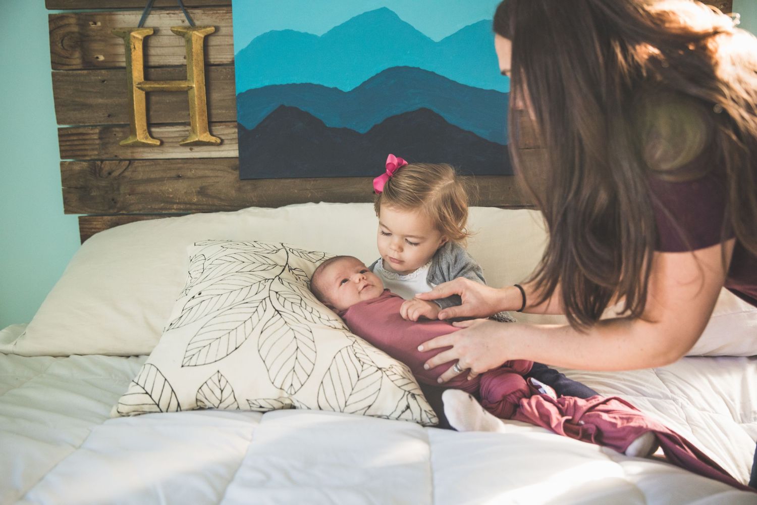 Siblings share tender moment with newborn baby on bed with mountain mural backdrop.