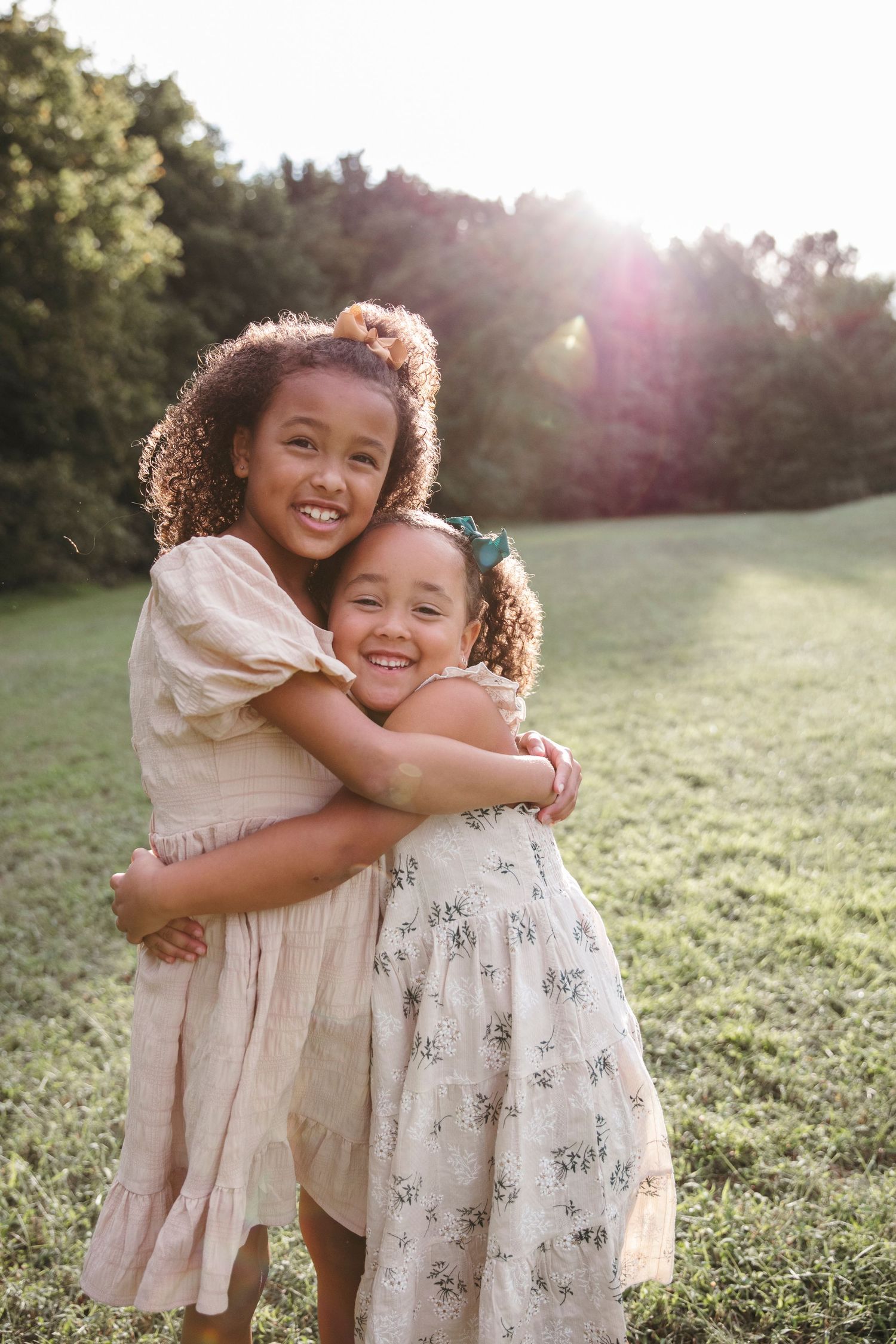 Two young children in light dresses share a joyful hug while standing in a sunlit grassy field.