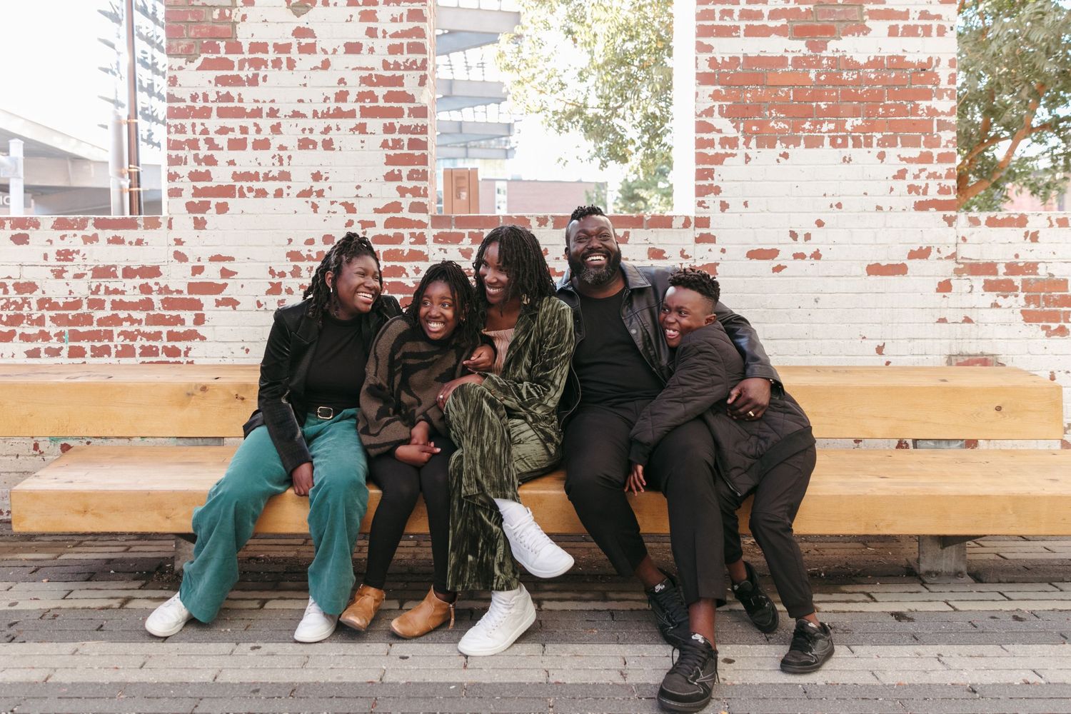 A family with middle school age children sitting together on wooden bench against rustic brick wall outdoors.