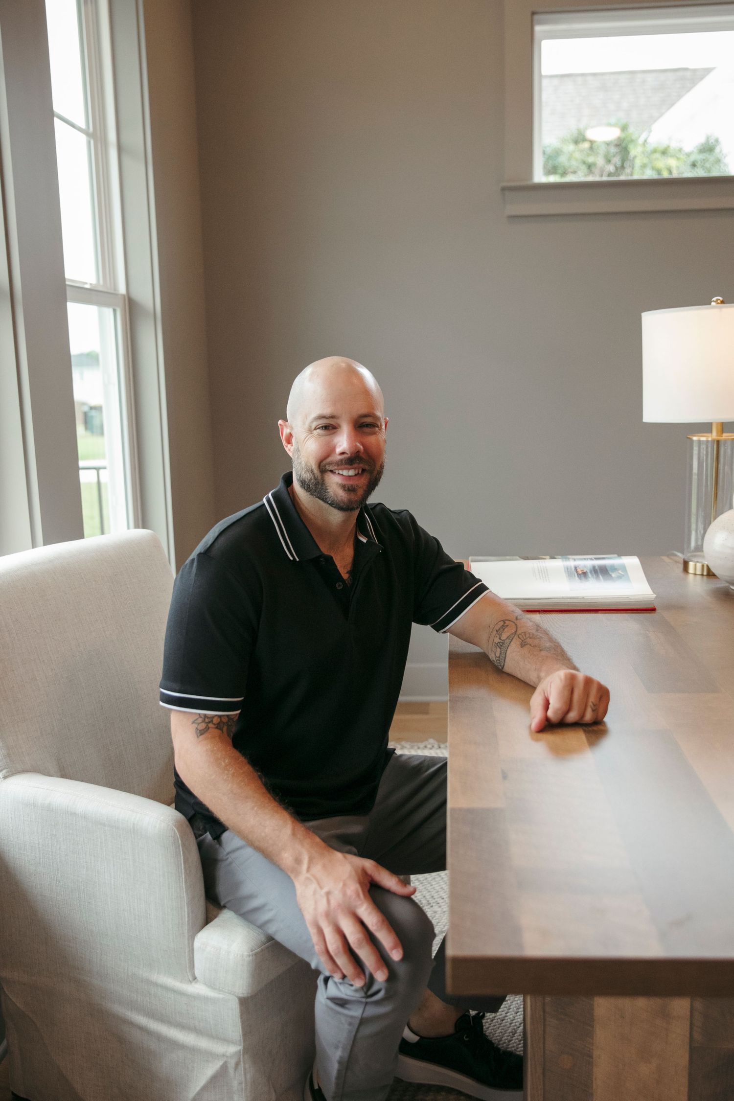 Professional headshot of person in black polo shirt seated at wooden desk near window.
