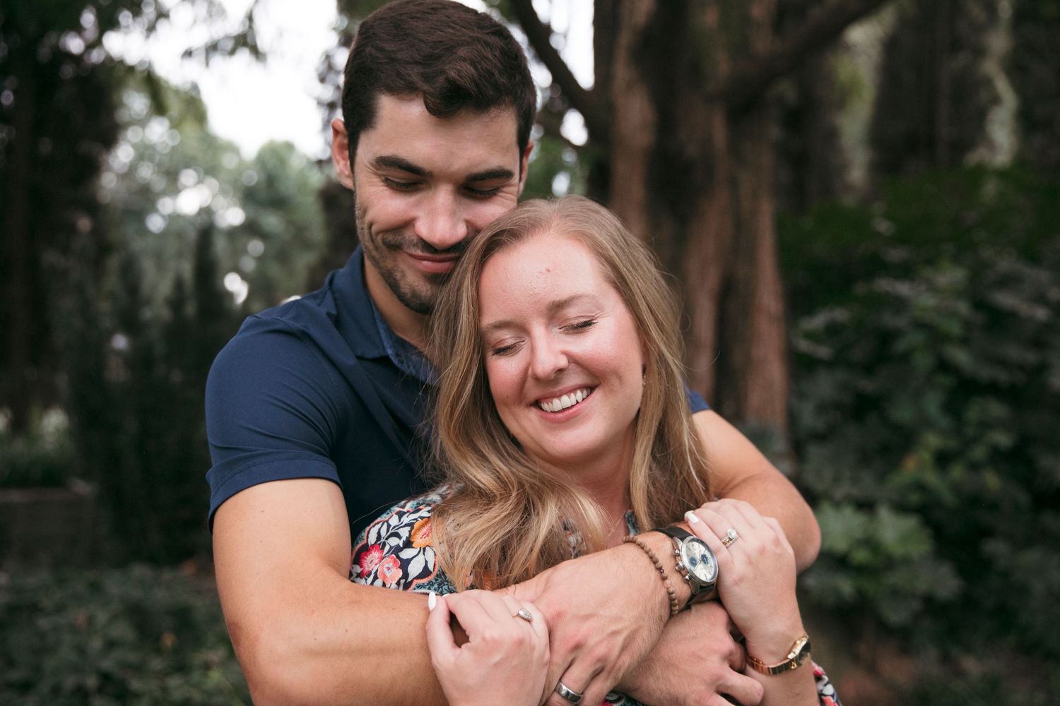A romantic couple embraces while smiling in a wooded outdoor setting.