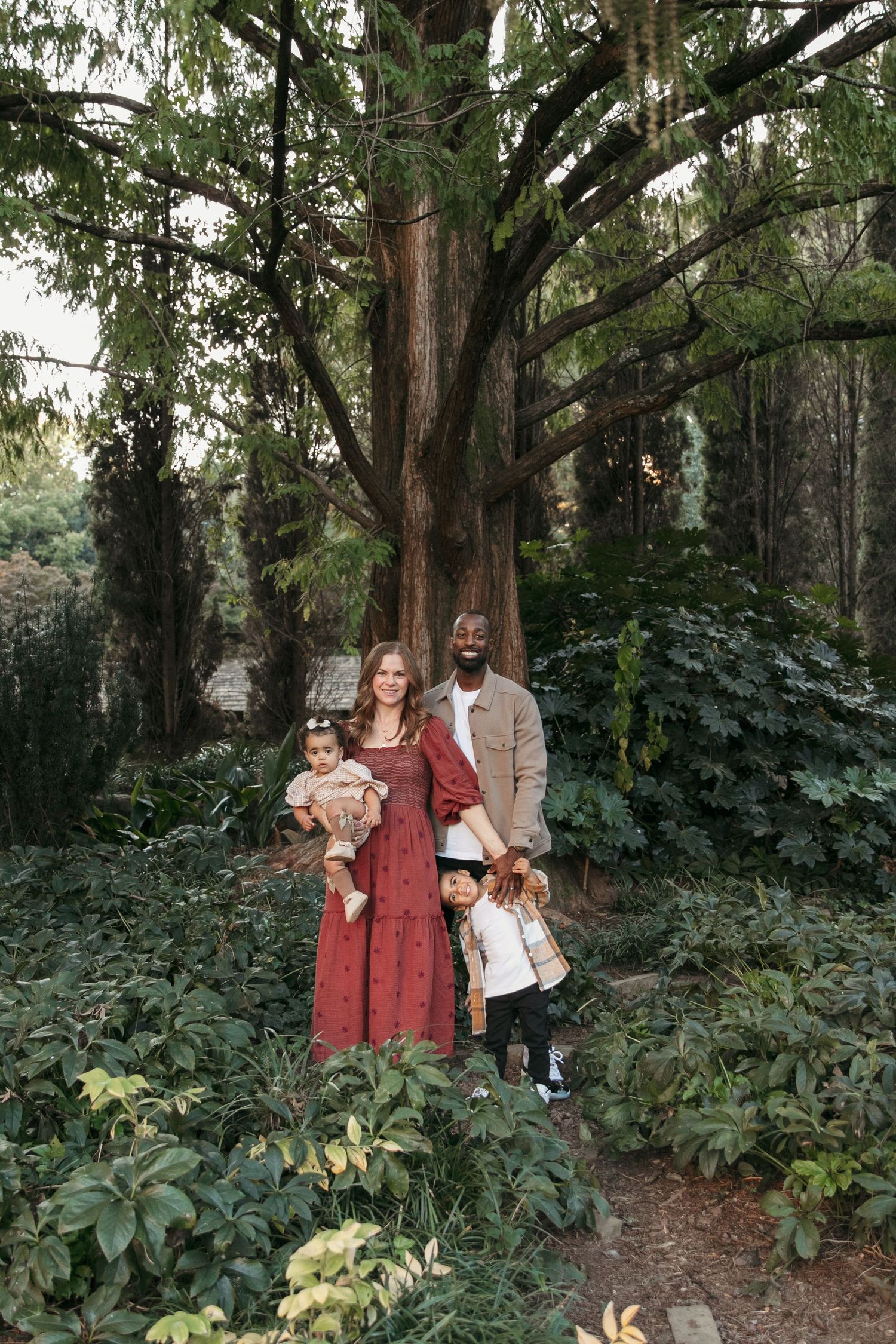 A family poses together under a majestic tree in a lush garden setting.