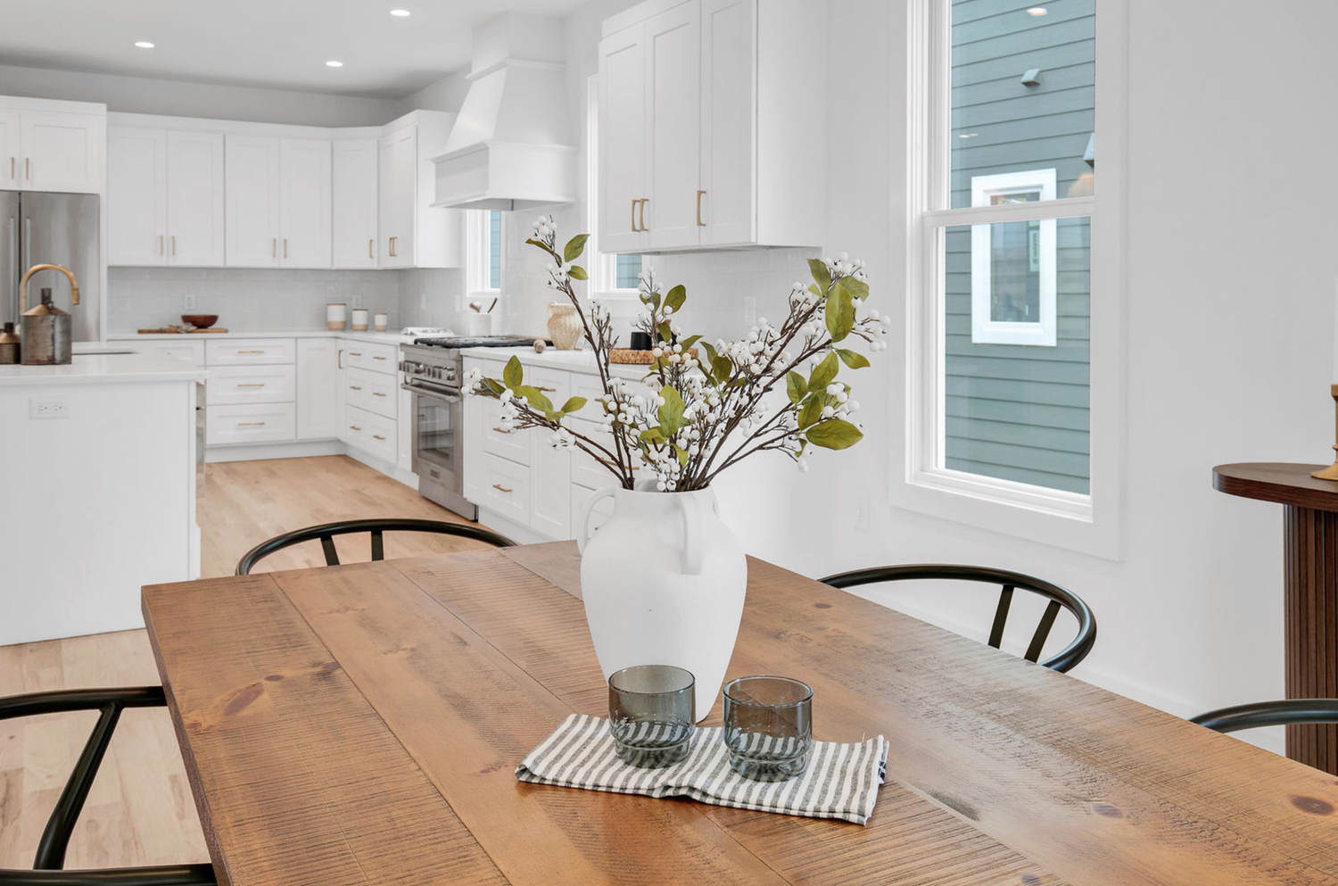 Bright dining area with rustic wood table, black chairs, white vase with branches, and modern white kitchen in background.