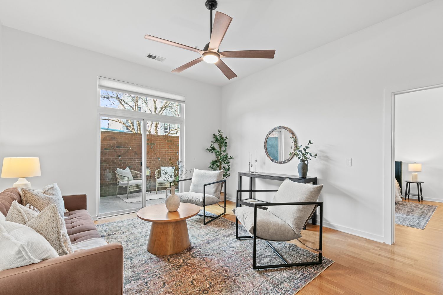 Bright modern living room with hardwood floors, leather sofa, ceiling fan, and large window overlooking a patio.