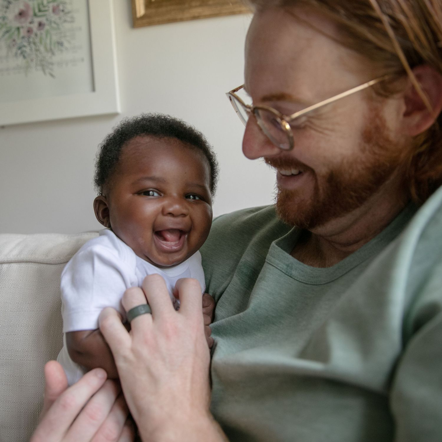 A joyful baby wearing white laughs while being held in a green shirt against a light interior background.