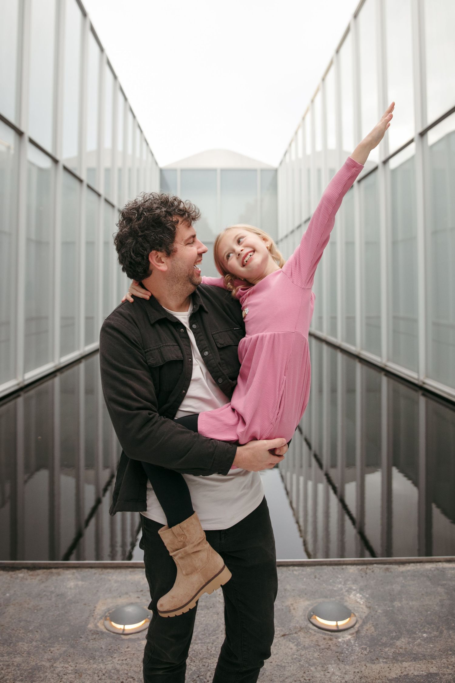 Adult holding child in pink dress who stretches arms playfully in modern corridor.