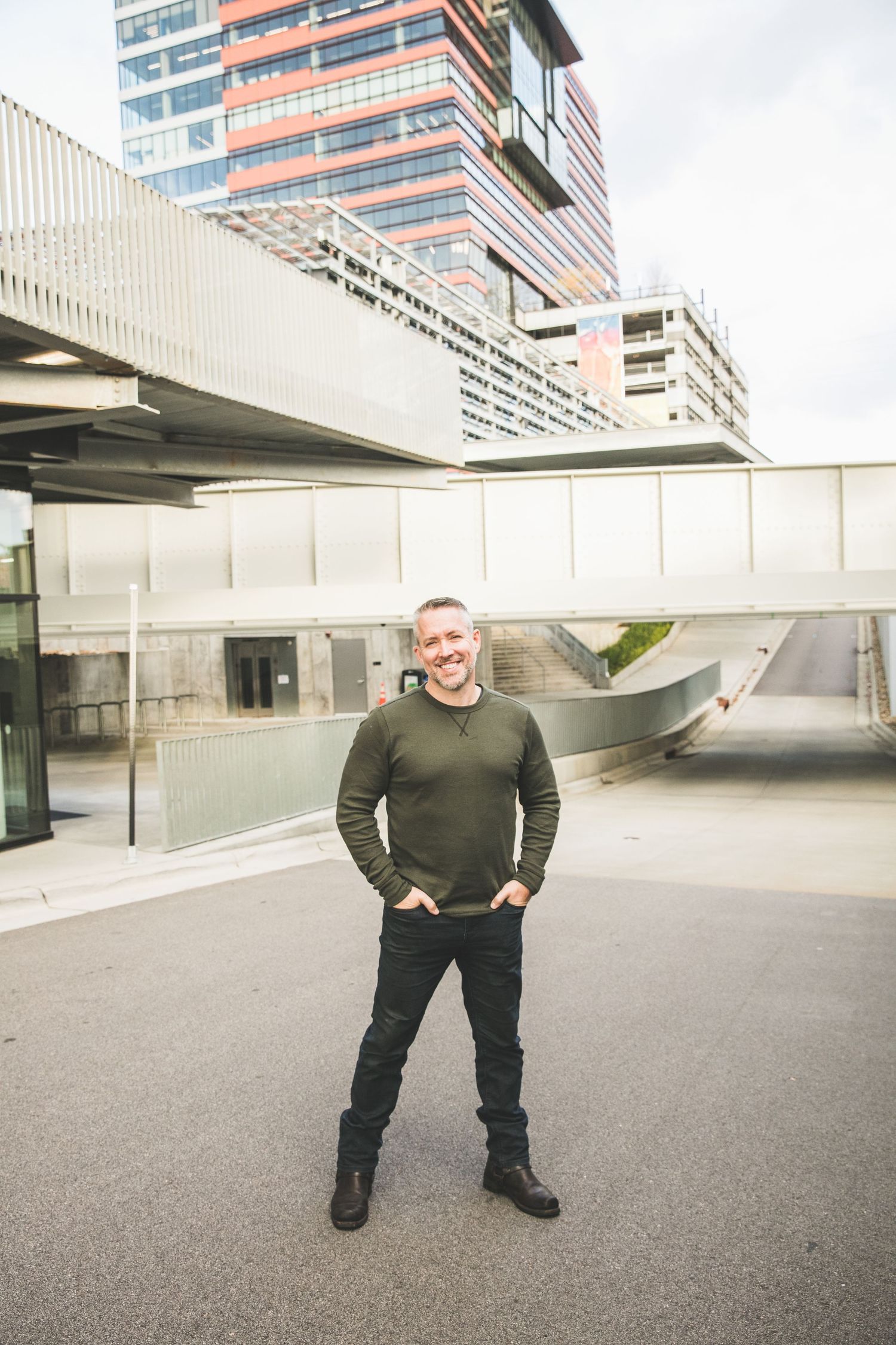 Person in olive sweater poses confidently against modern architectural backdrop.
