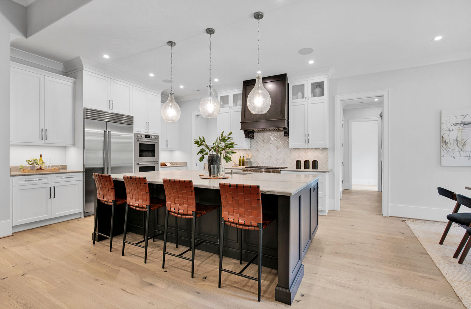Modern kitchen with white cabinets, large island, leather bar stools, pendant lights, and stainless steel appliances on light hardwood floor