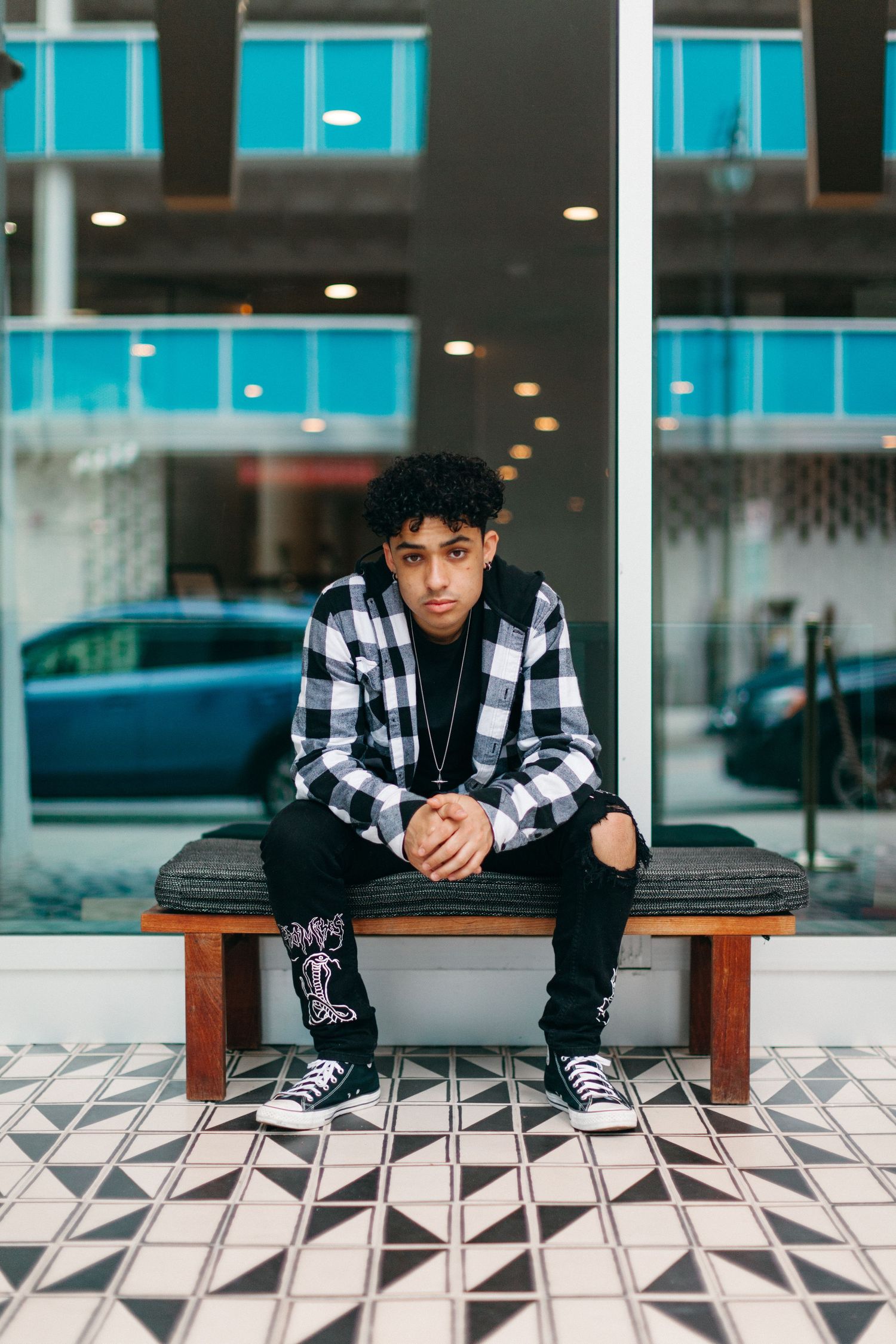 A person in a black and white plaid shirt sits on a wooden bench against a modern building with geometric floor tiles.