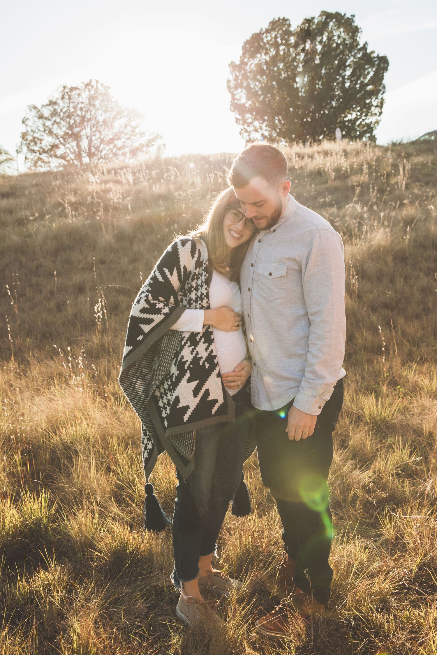 Pregnant couple sharing intimate moment in sunlit field with tall grass at golden hour.
