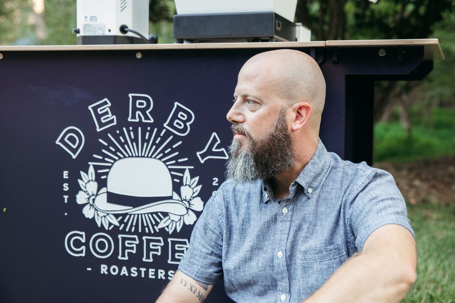 Coffee roaster with beard wearing grey shirt next to Derby Coffee cart outdoor display.