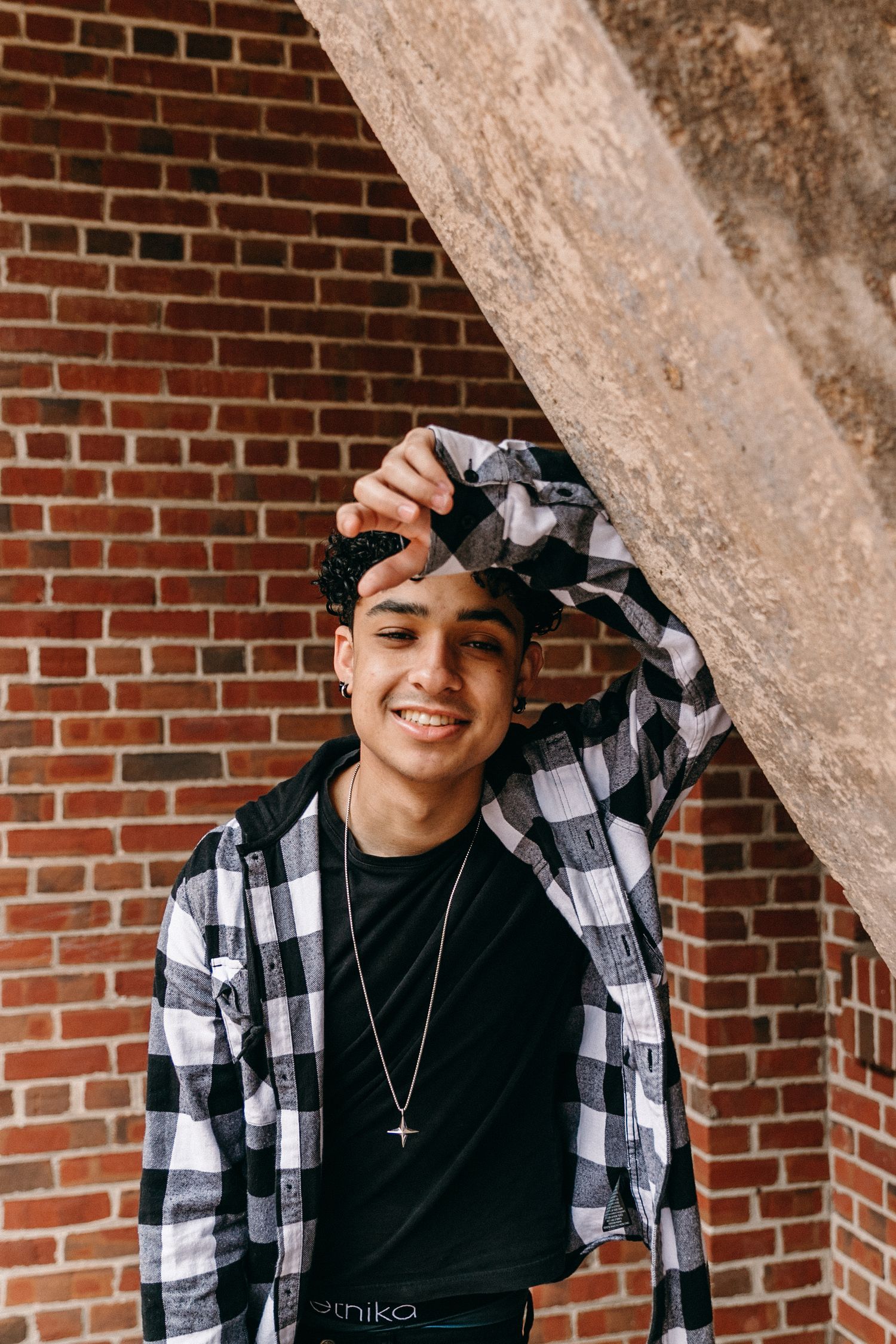 Person wearing black and white plaid flannel and necklace against brick wall background.