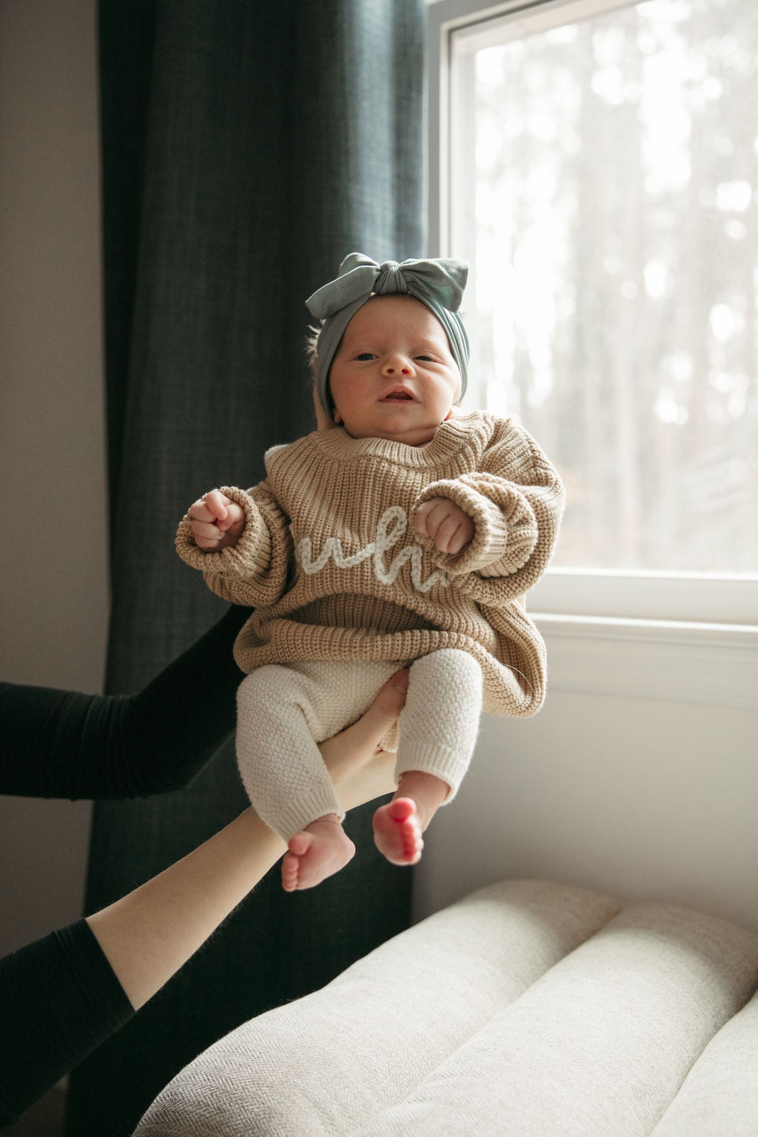 A baby wearing a cozy beige knit sweater lies peacefully near a bright window in a lifestyle photography series.