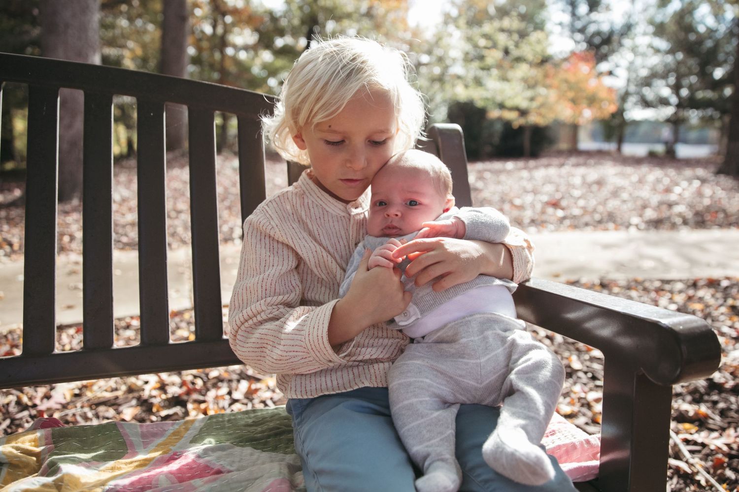 A baby in light colored clothing sits with someone on a wooden bench surrounded by fallen leaves.