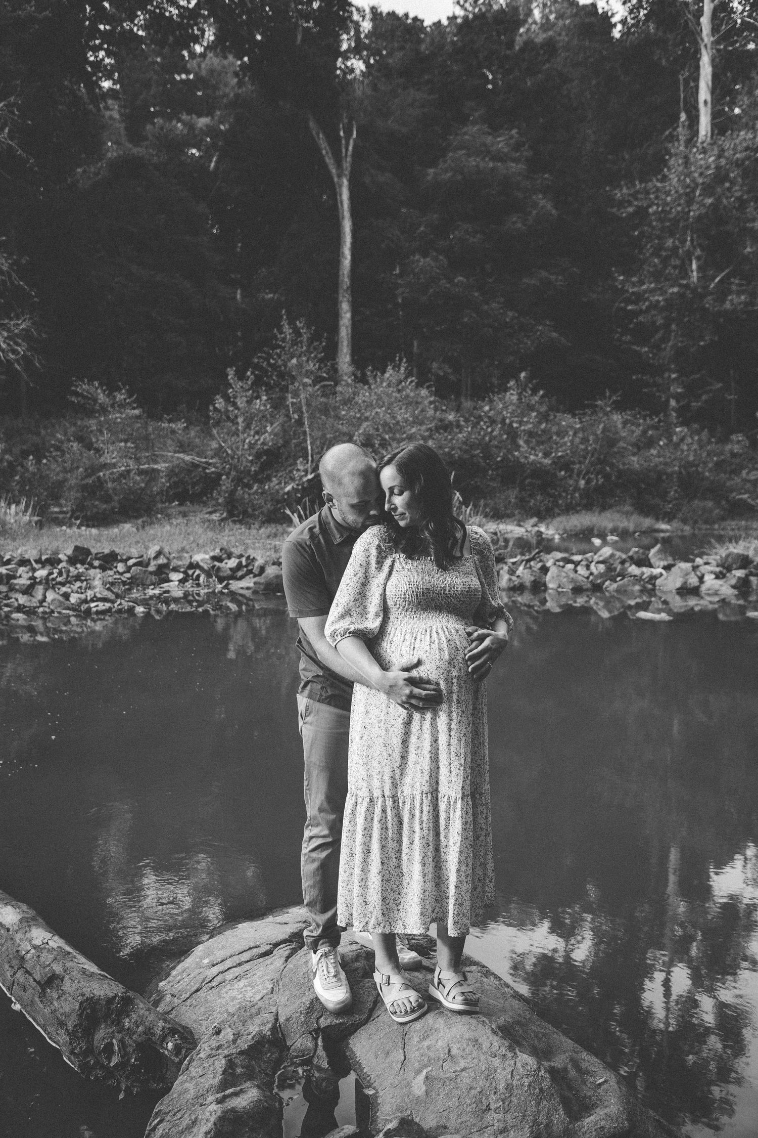 Maternity photo of a couple standing on rocks by a serene lake in black and white.